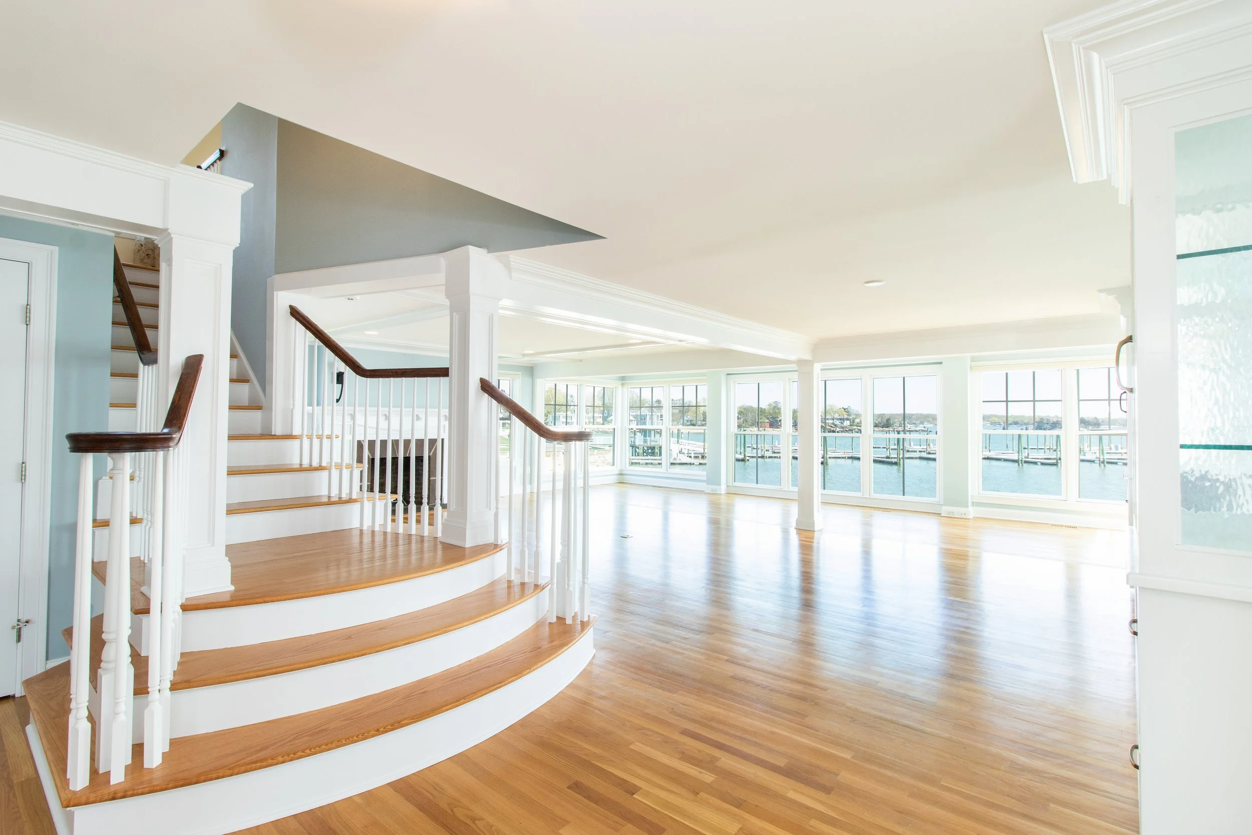 Bright, spacious living area with large windows overlooking water, light-colored walls, hardwood floors, and staircase with white railing and dark wooden handrail.