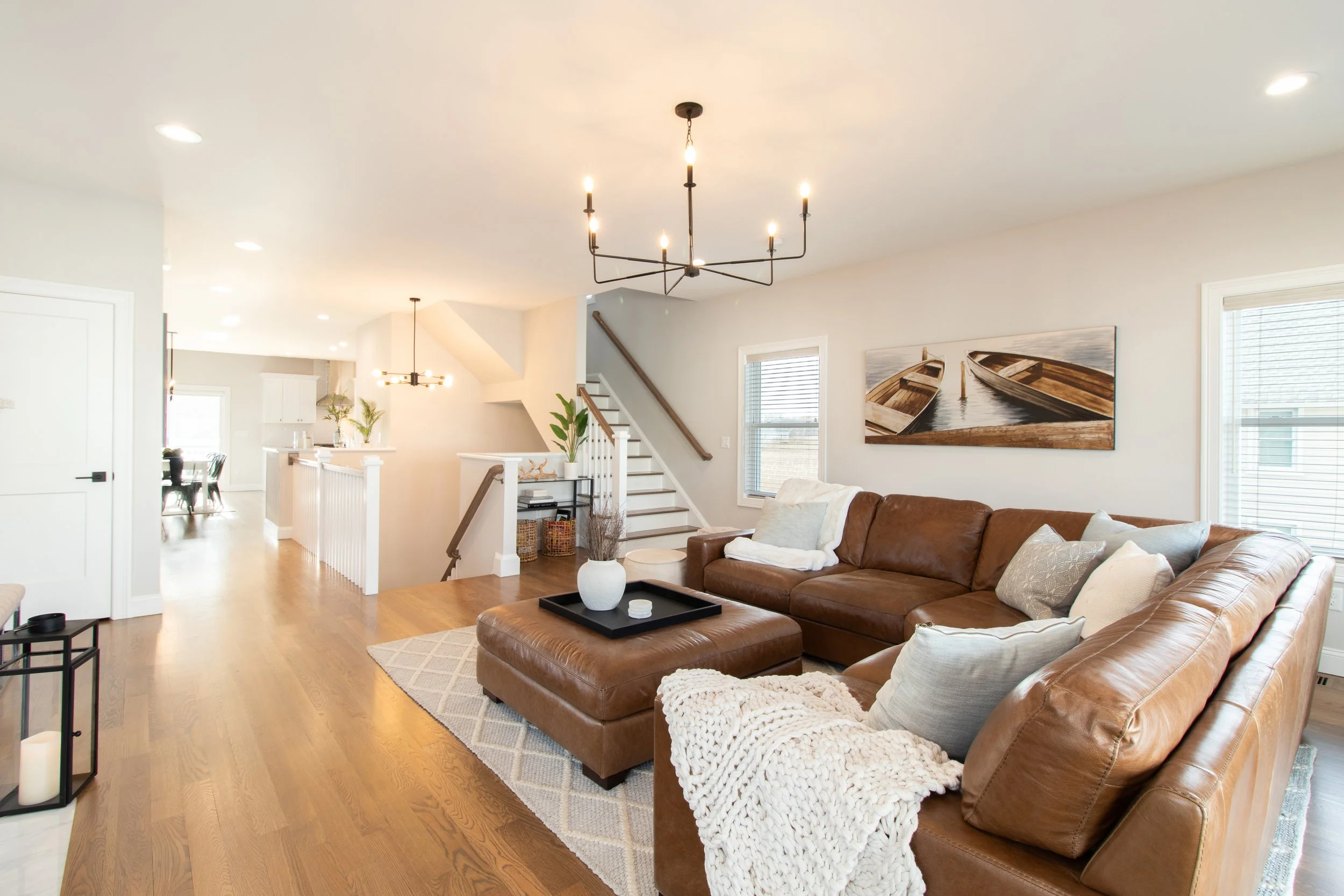 Living room with brown leather sectional sofa, wooden tray with decorative items, large wall art of boats, hardwood floors, and large windows with blinds.