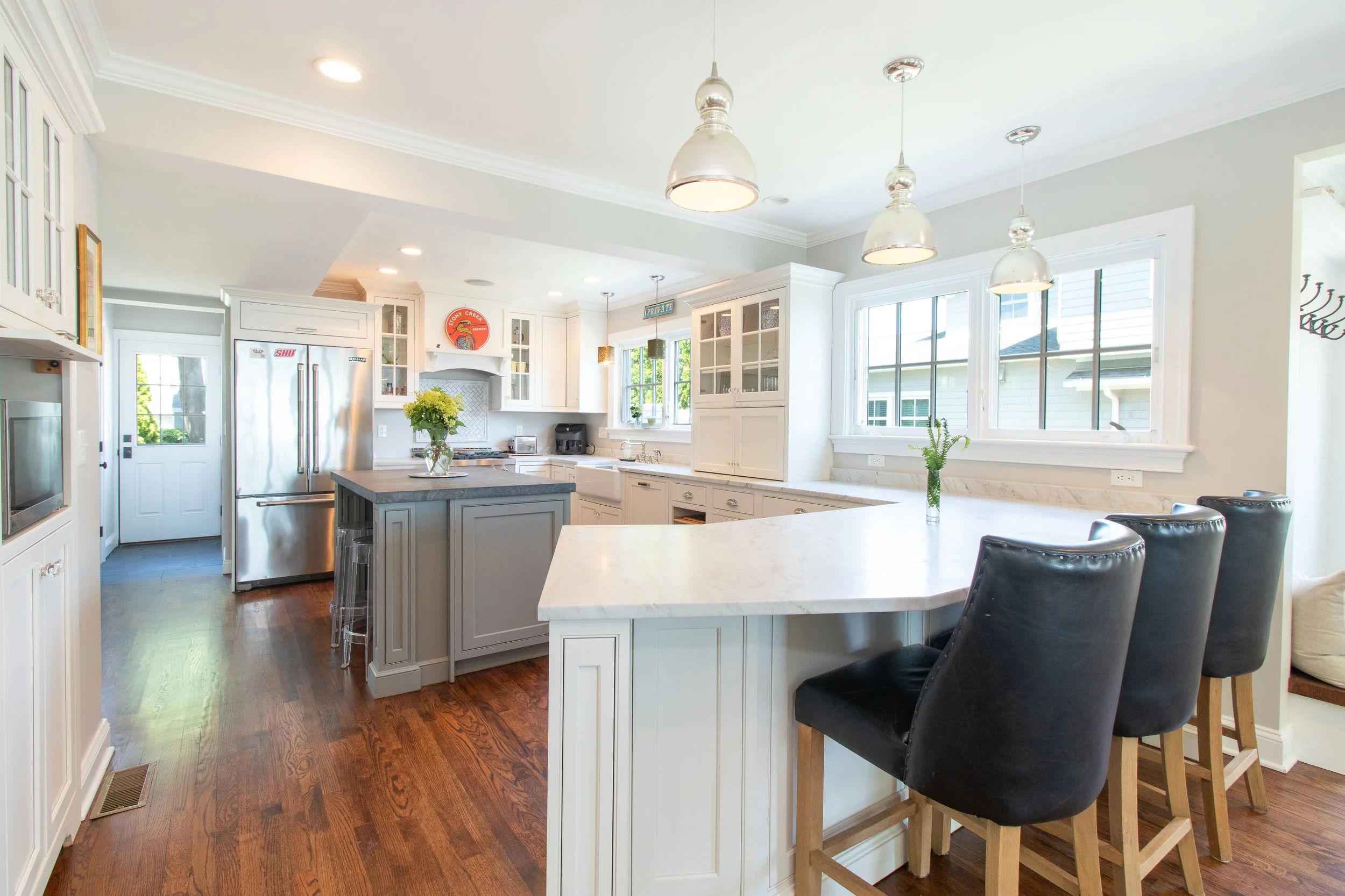 Bright, modern kitchen with white cabinets, a marble countertop breakfast bar, black leather chairs, wooden floors, and large windows letting in natural light.