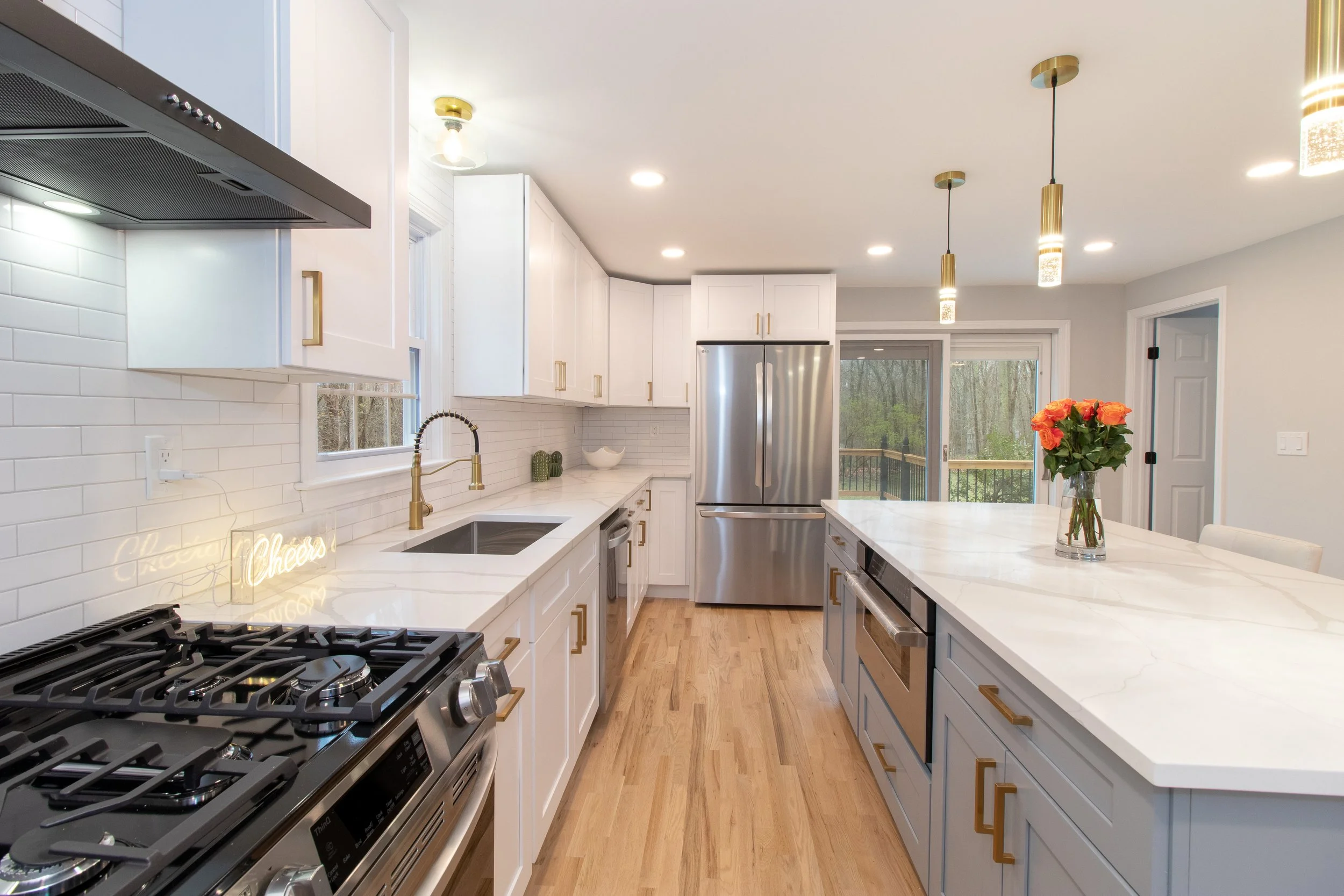 Modern kitchen with white cabinets, a large island with a marble countertop, stainless steel refrigerator, oven, and gas stovetop. Decor includes a vase of orange roses and a neon sign with the word Cheers.