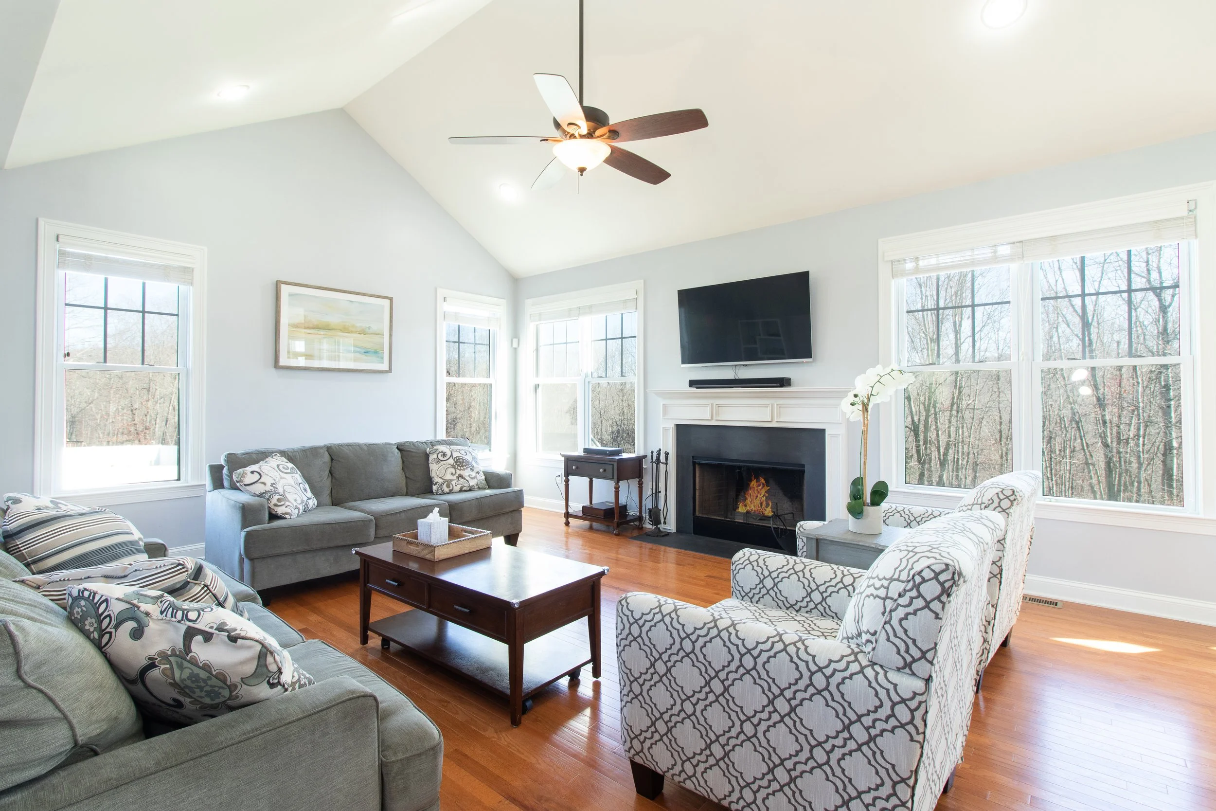 Bright living room with light gray walls, hardwood floors, a fireplace with a mounted TV above, and multiple windows letting in natural light. The room has a gray sofa, patterned armchairs, a wooden coffee table, a small side table, and a potted orch
