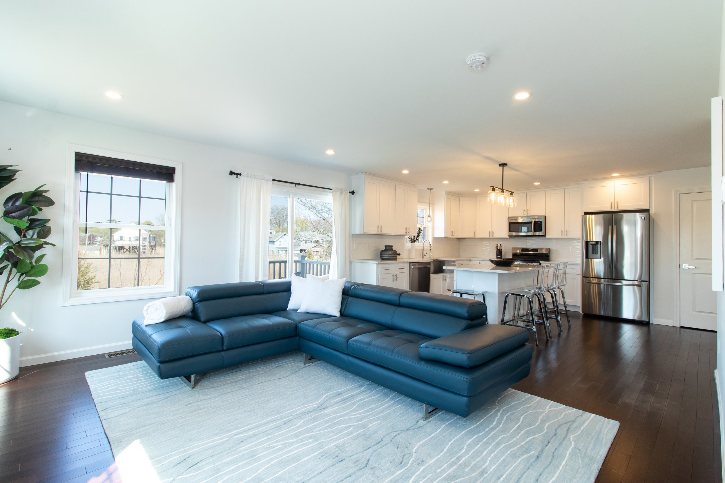 Living room with a dark blue sectional sofa, a white area rug, and large windows with white curtains, opening to a view of neighboring houses, adjacent to a modern white kitchen with stainless steel appliances.