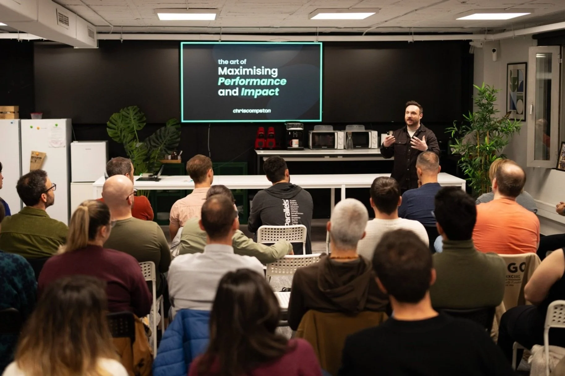 Chris Compston giving a presentation in front of an audience, with a screen displaying the title 'The Art of Maximising Performance and Impact.' The speaker is in a room with chairs and a table, surrounded by attendees seated and listening.