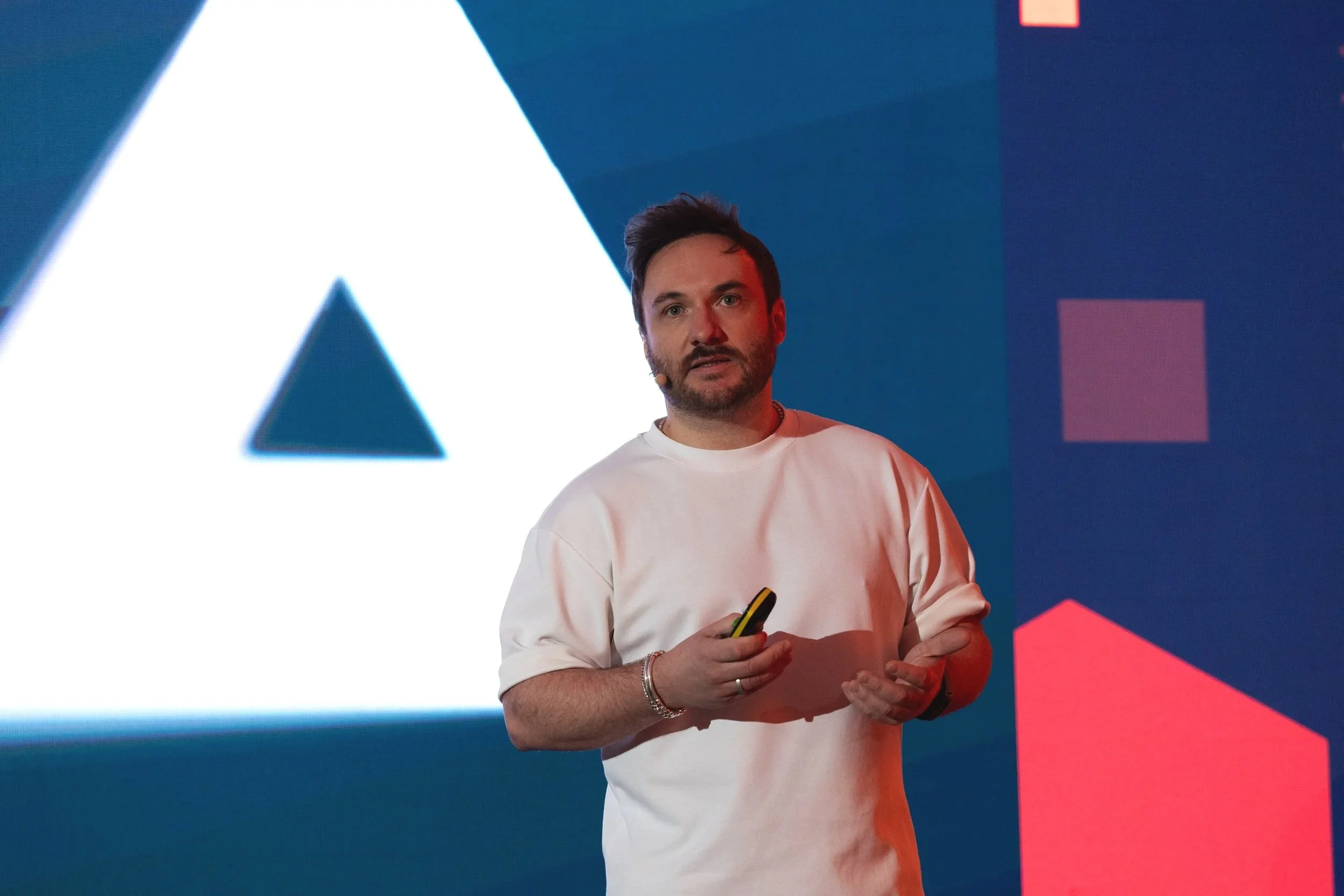 Chris Compston, holds a remote control while standing on a stage with large graphics behind him. The background features a large white triangle with a smaller blue triangle inside it on the left, and geometric shapes in pink and blue on the right.