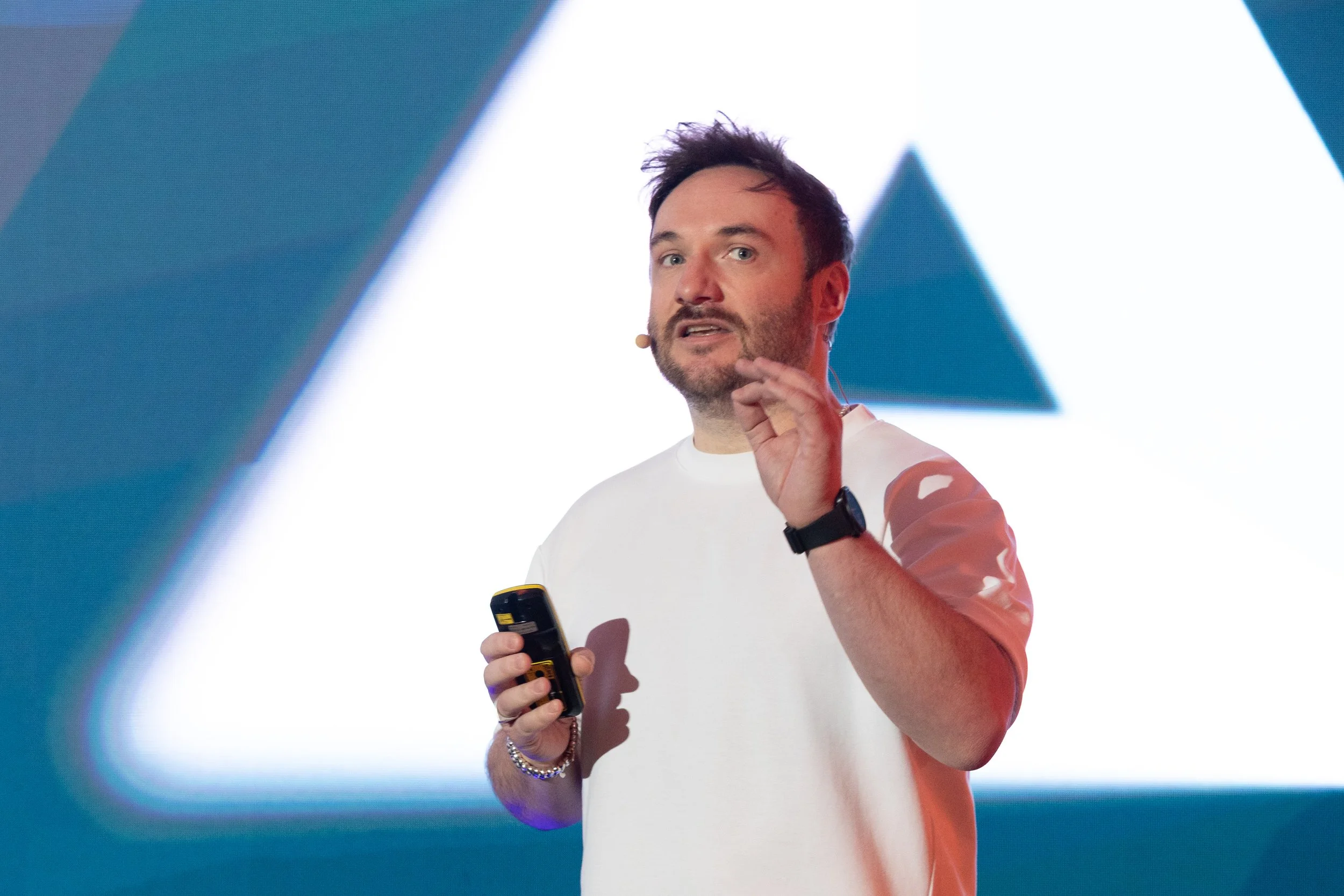 Chris Compston, wearing a white t-shirt, is holding a remote control in his right hand and gesturing with his left hand during a presentation. Behind him is a large screen displaying geometric shapes.