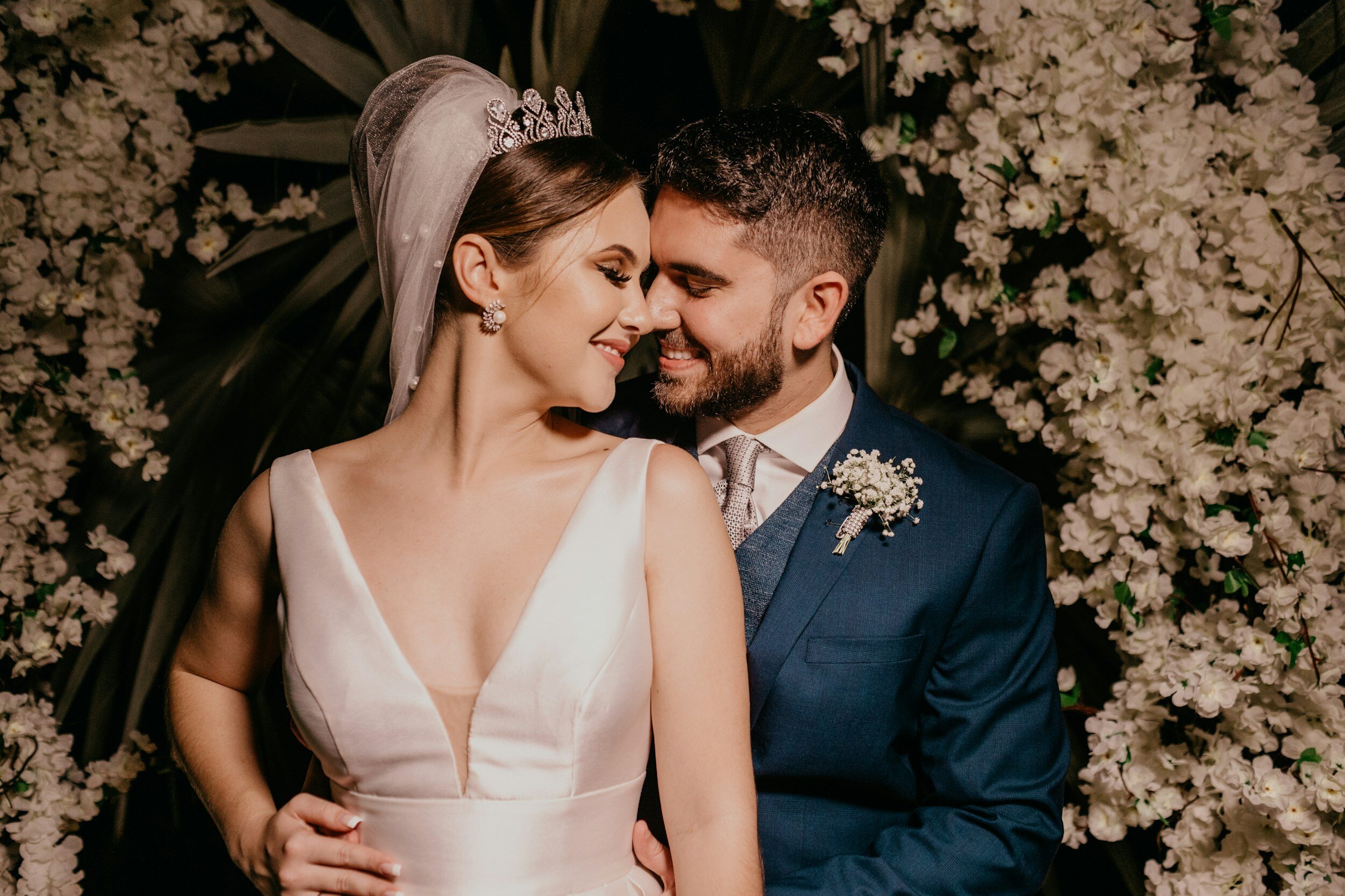 A bride and groom sharing a close, intimate moment at their wedding, surrounded by white flowers. The bride is wearing a white wedding dress with a deep V neckline, a veil, and a tiara, while the groom is dressed in a blue suit with a white shirt, a tie, and a floral boutonniere.