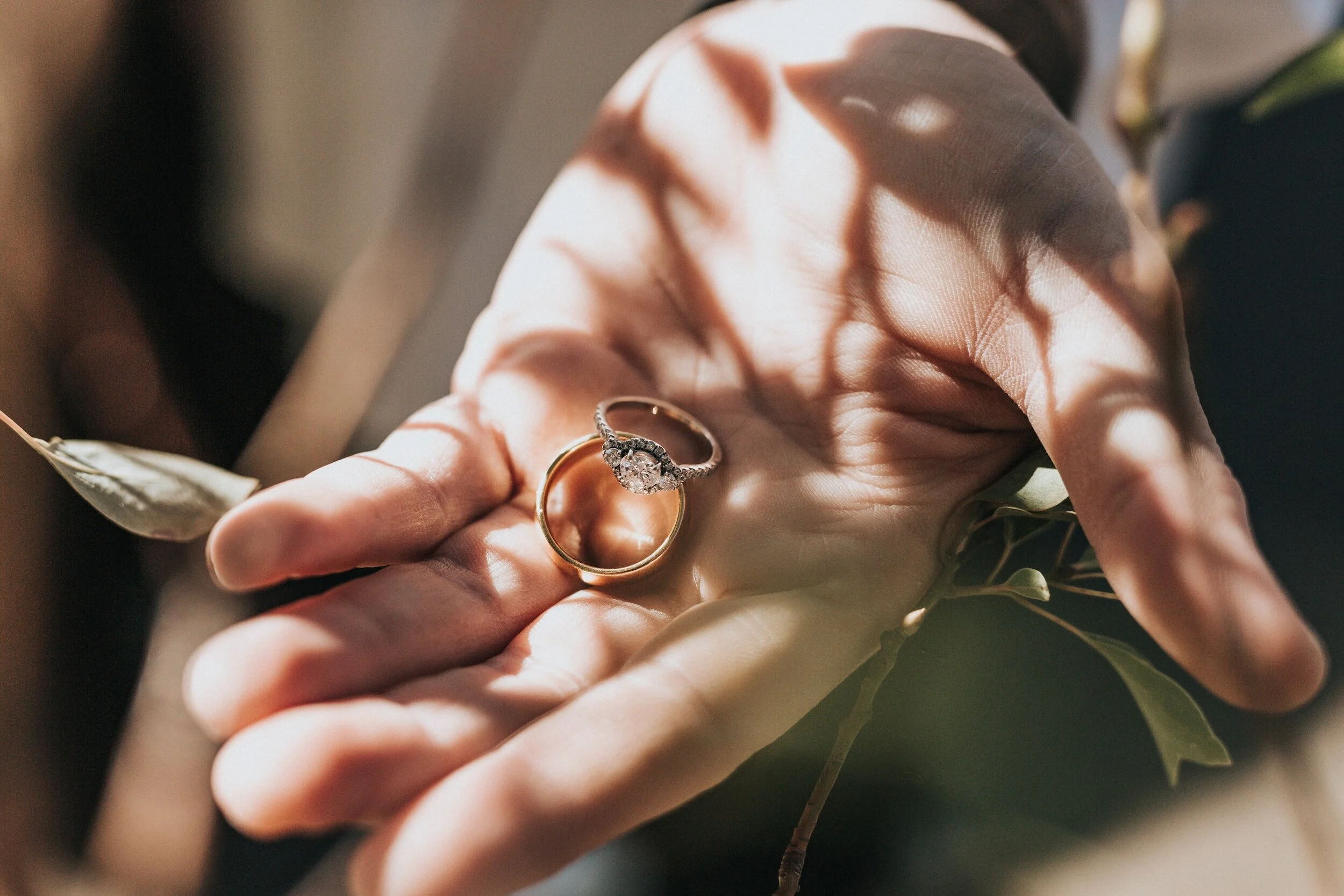 A hand holding two rings, one with a diamond engagement ring design and the other a plain gold band, with soft lighting and shadows of leaves on the hand.