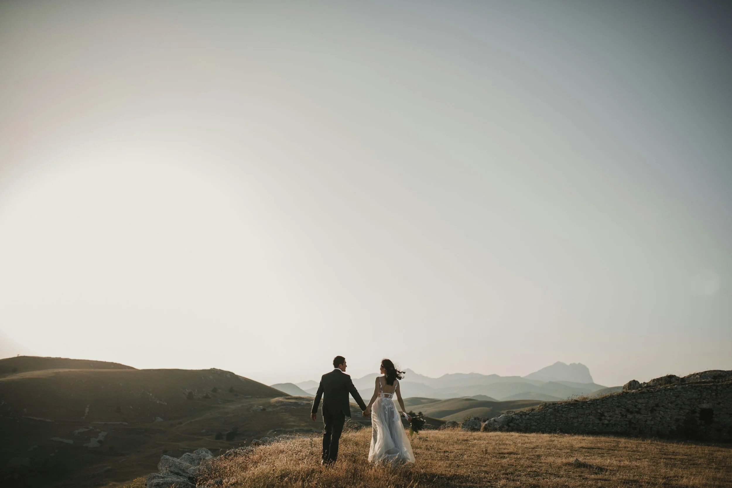A couple dressed in wedding attire walking hand-in-hand on a grassy hilltop at sunset, with mountains in the background.