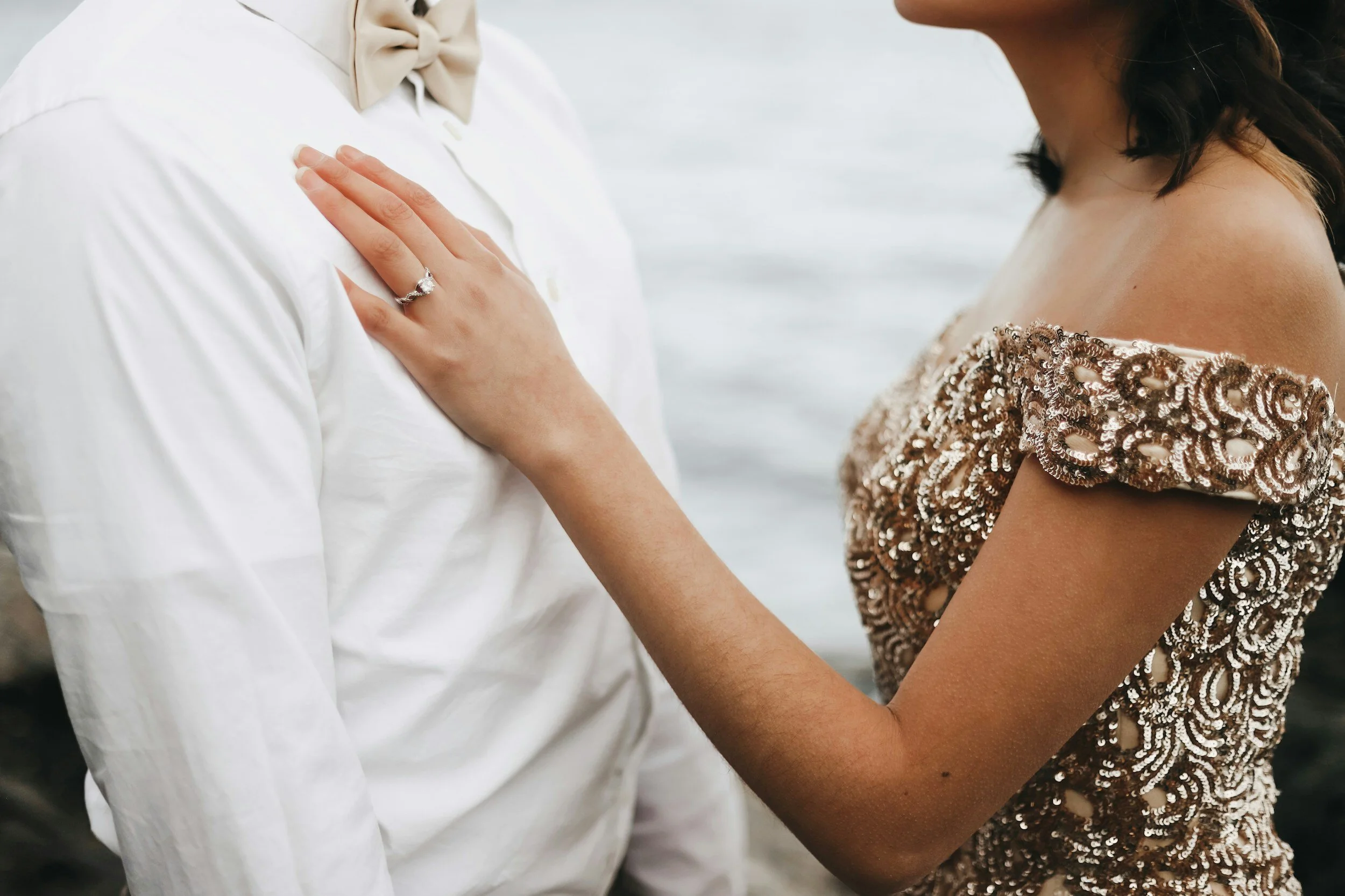 A woman in a sequined off-shoulder gown and a man in a white shirt with a bow tie are close together outdoors near water, with the woman's hand on the man's chest showing a diamond ring.