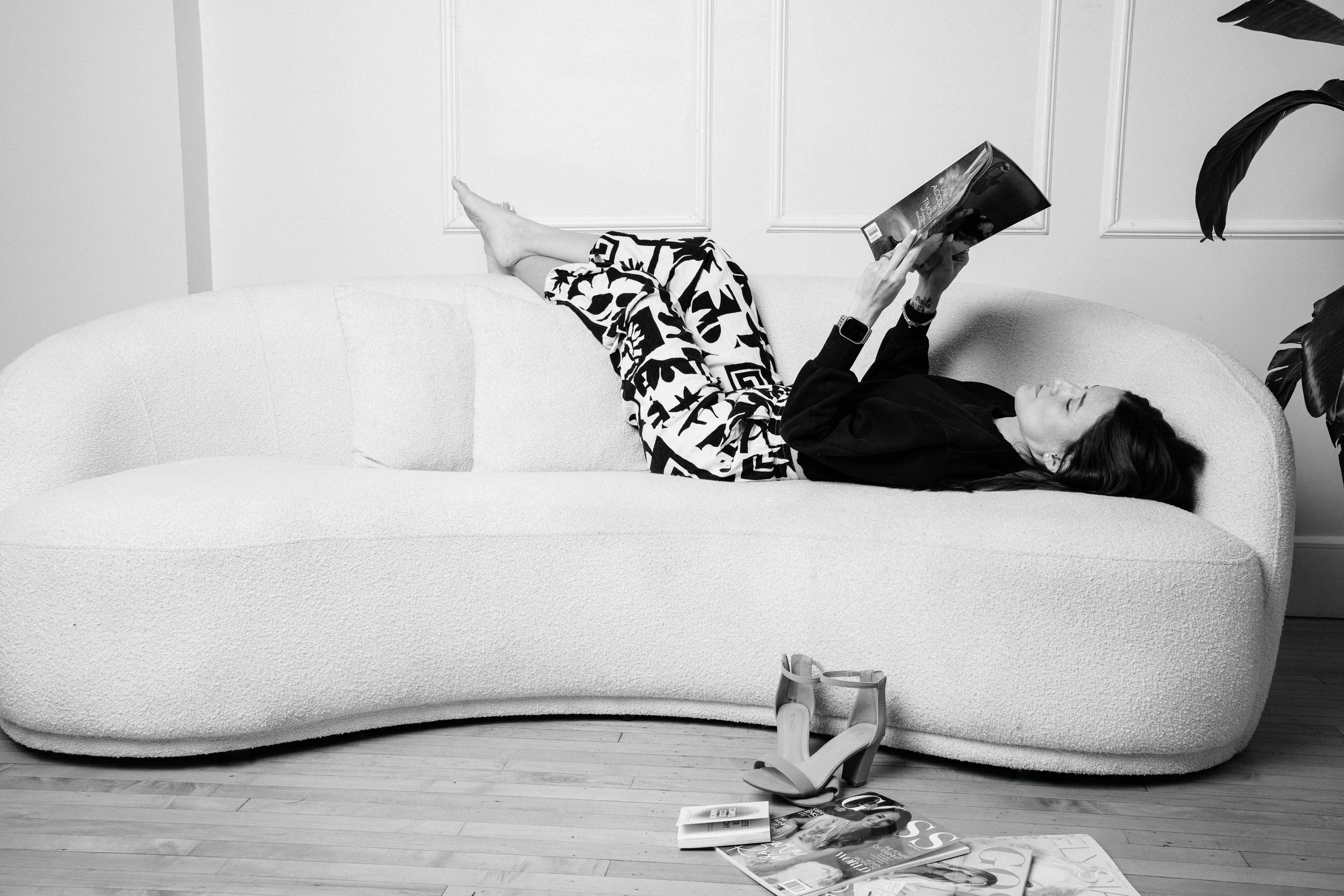 A woman lying on a modern white curved sofa, reading a book, with high-heeled shoes and magazines on the floor nearby.
