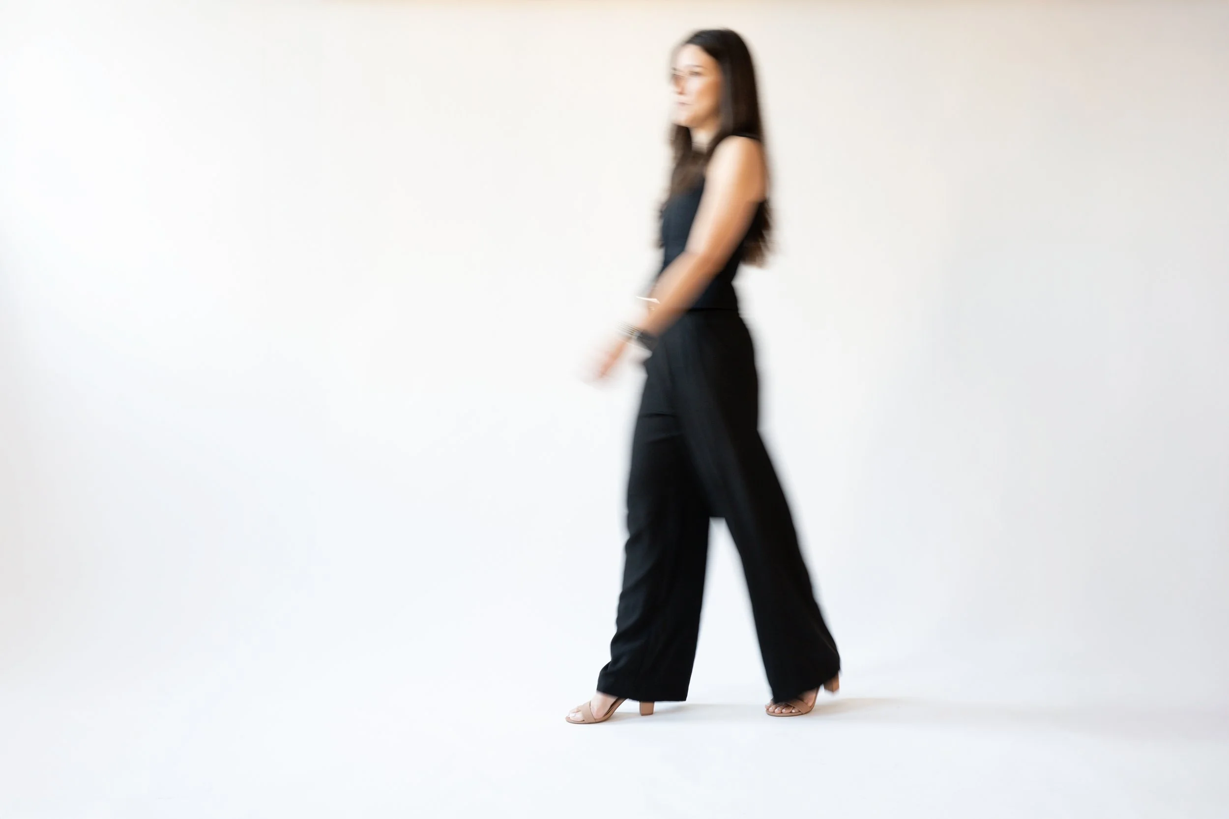 A woman in black clothing and heels is walking, with a white background.