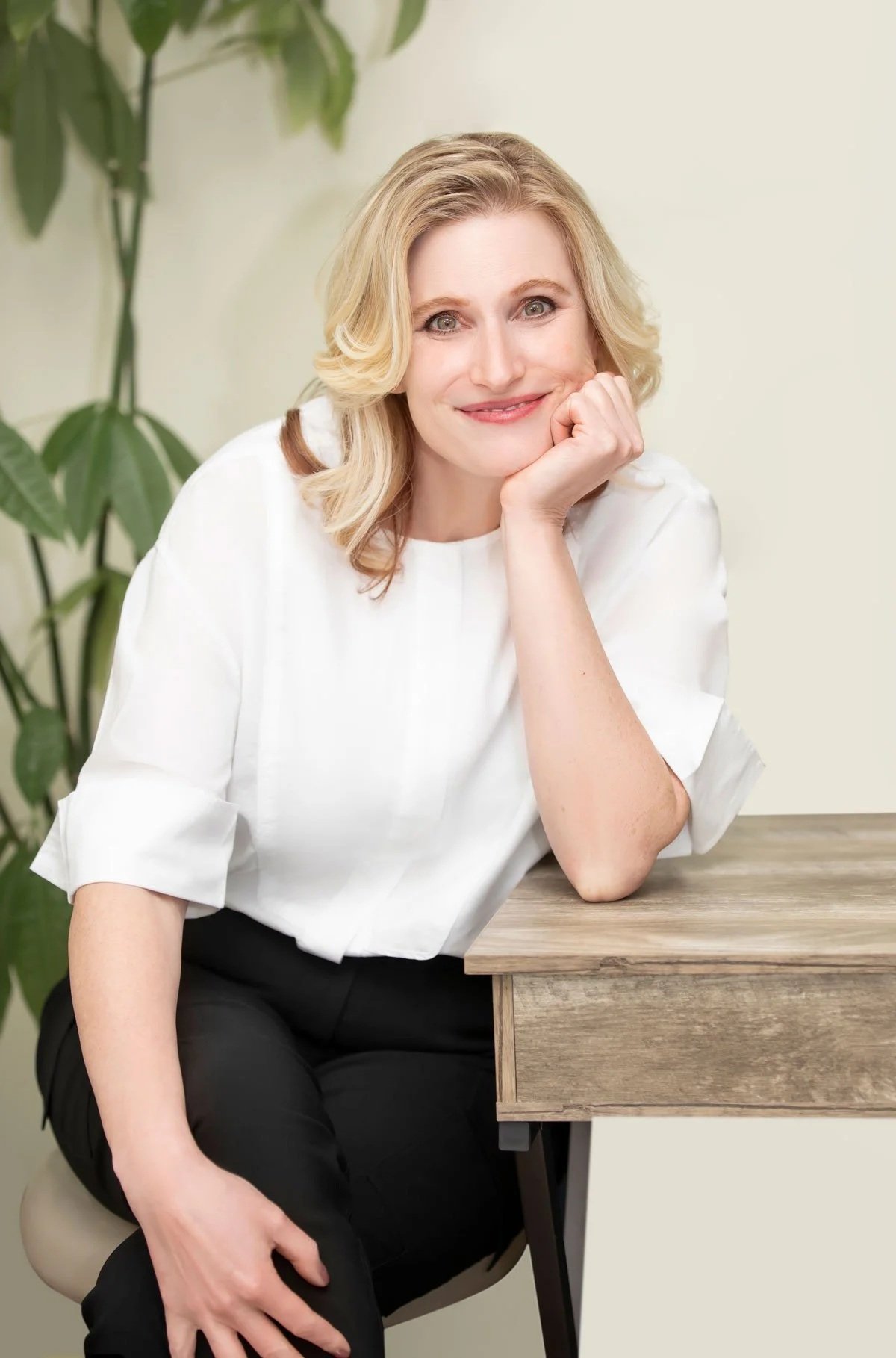 A woman with blonde hair, wearing a white blouse, sitting at a desk, smiling, leaning her chin on her hand, with a green plant in the background.