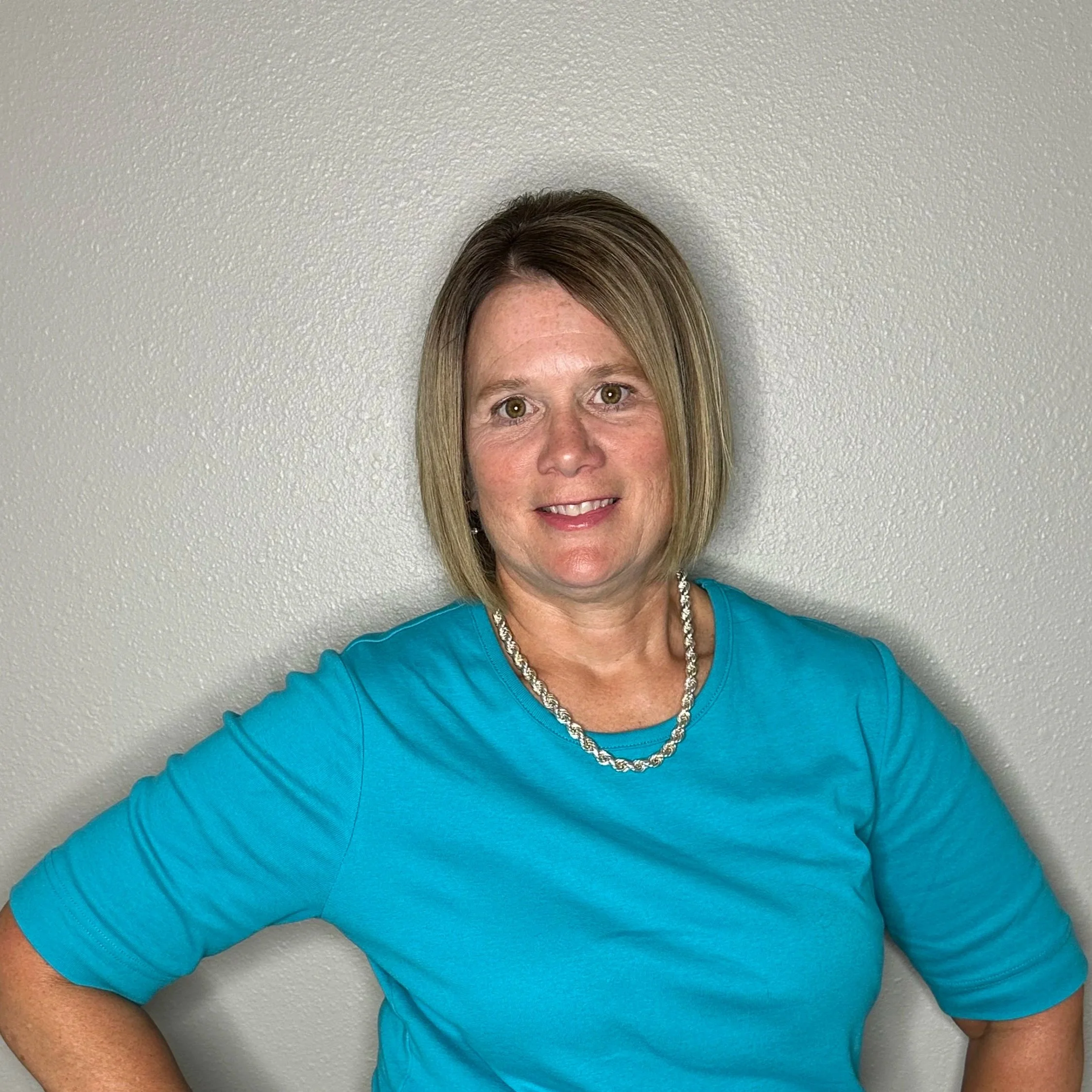 A woman with short, light brown hair wearing a bright blue t-shirt, a silver necklace, and a smartwatch standing against a plain light gray wall. She has her hands on her hips and is smiling at the camera.