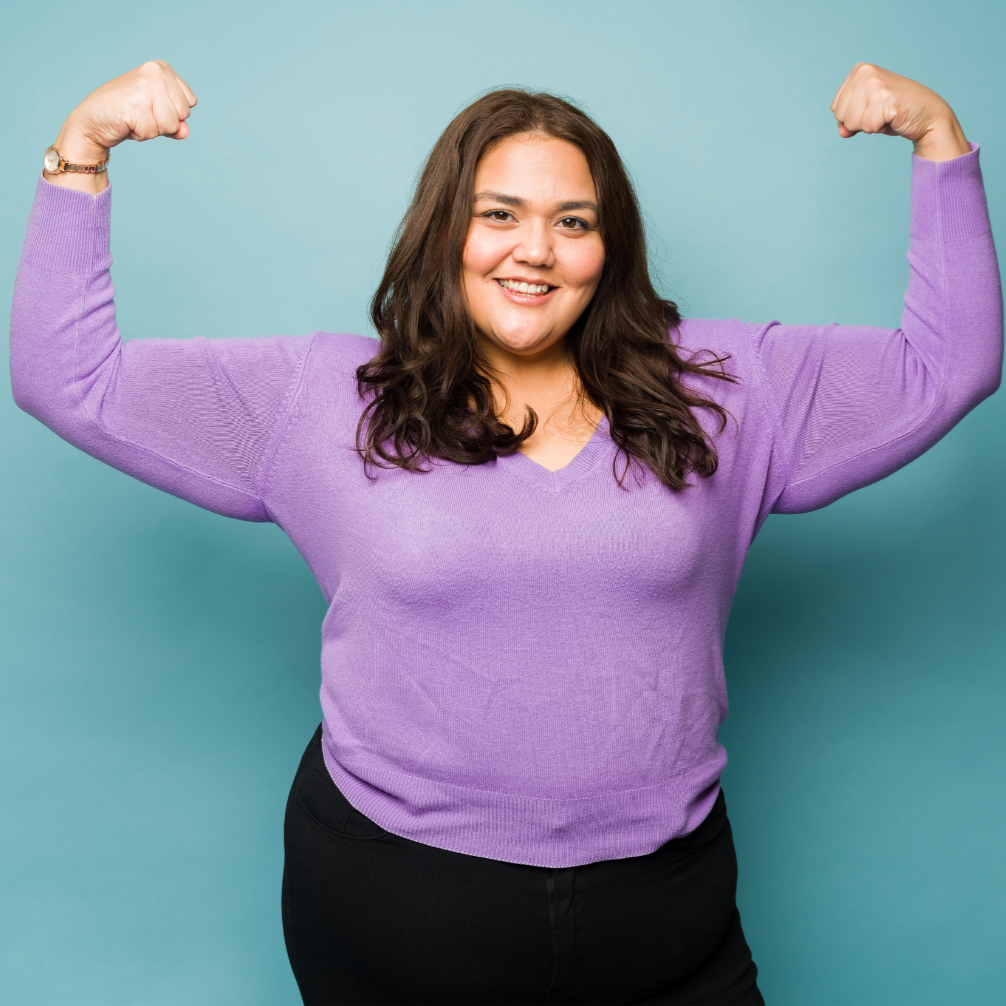 A smiling woman with brown hair and a purple sweater flexing her biceps in front of a light blue background.