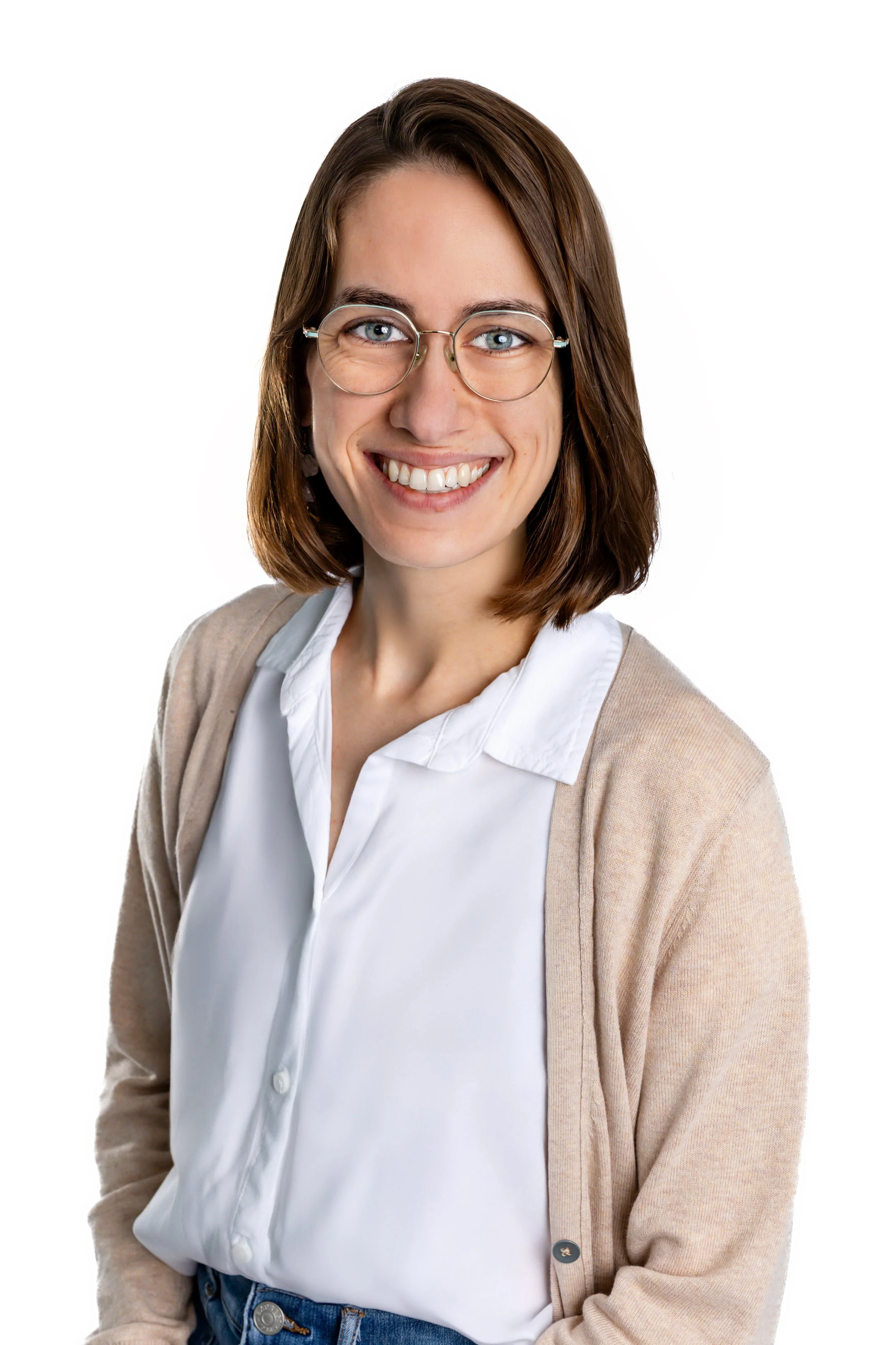 A woman with brown hair and glasses sitting on a beige armchair smiling at the camera, wearing a white shirt, beige cardigan, blue jeans, and beige sneakers, in a room with light blue walls, a blue side table, and a floor lamp.