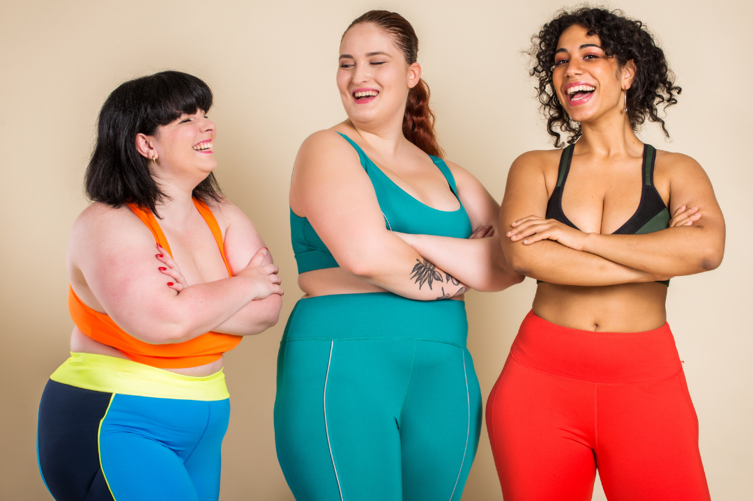 Three women in colorful workout clothes laughing and smiling together against a beige wall.