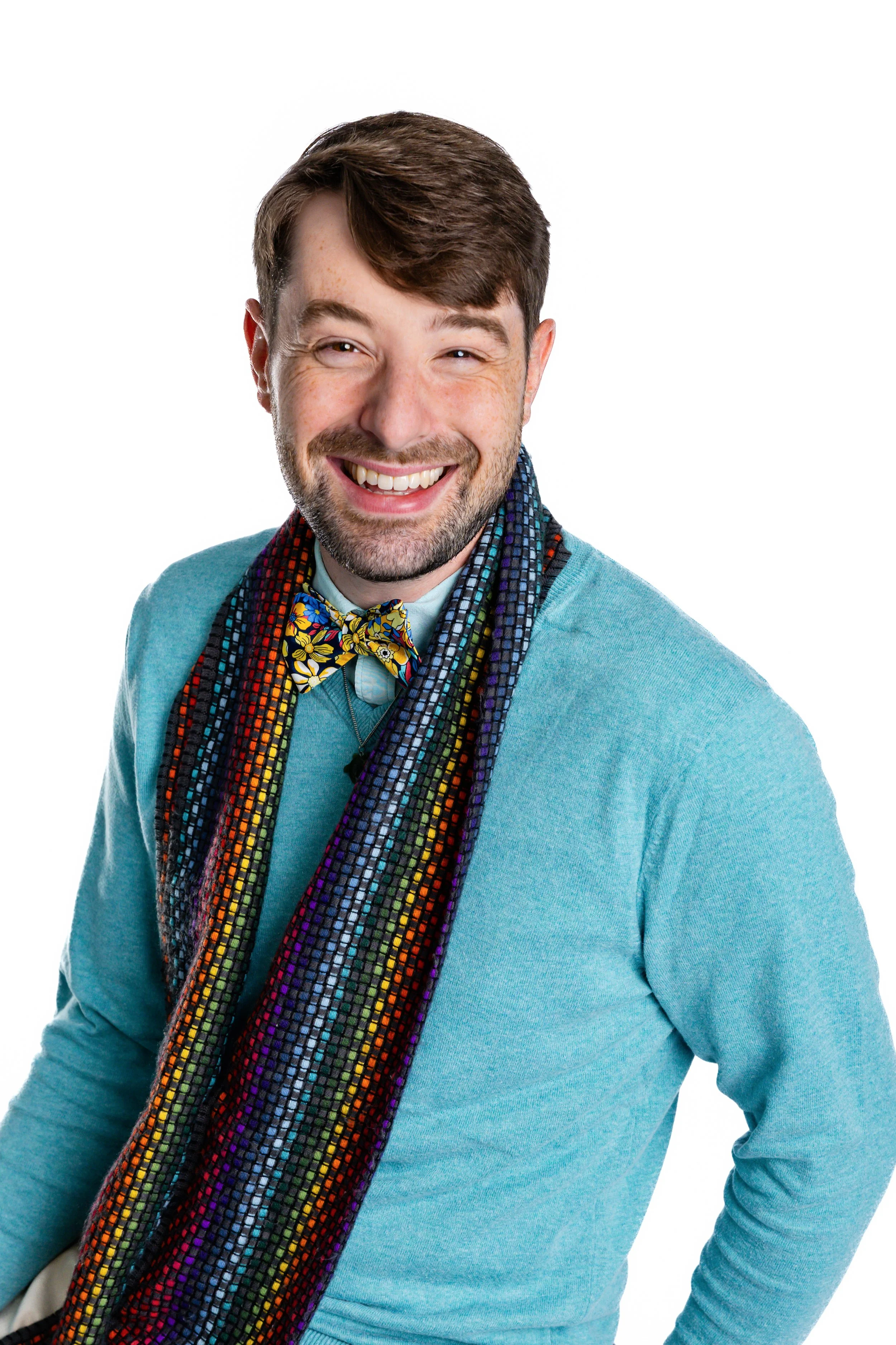 A man with brown hair and a beard, wearing a light blue sweater, beige pants, a colorful scarf, and multicolored checkered sneakers, sits on a beige armchair smiling at the camera. He is in a cozy room with blue walls, a table with a lamp, and decorative items including a sign that says 'Go with the waves.'