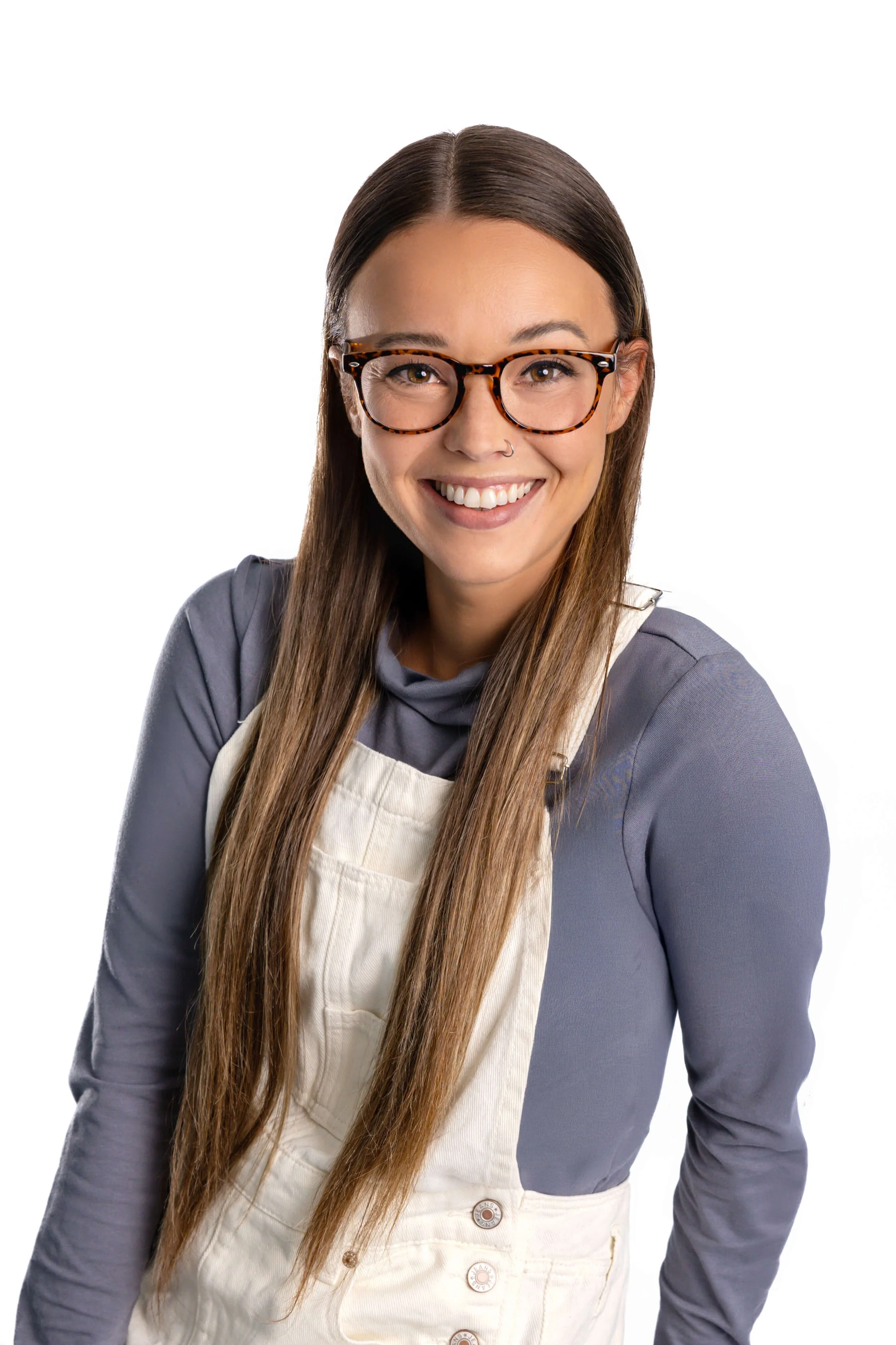 A young woman with long brown hair and glasses, sitting on a beige armchair, smiling at the camera. She's wearing a gray long-sleeve shirt under a white jumpsuit, with bracelets on her wrist and brown sandals. To her right, there is a wooden side table with a wicker lamp and a sign that reads "Go with the Waves." The background features a light blue wall, a blue cabinet with files underneath, and a standing lamp.