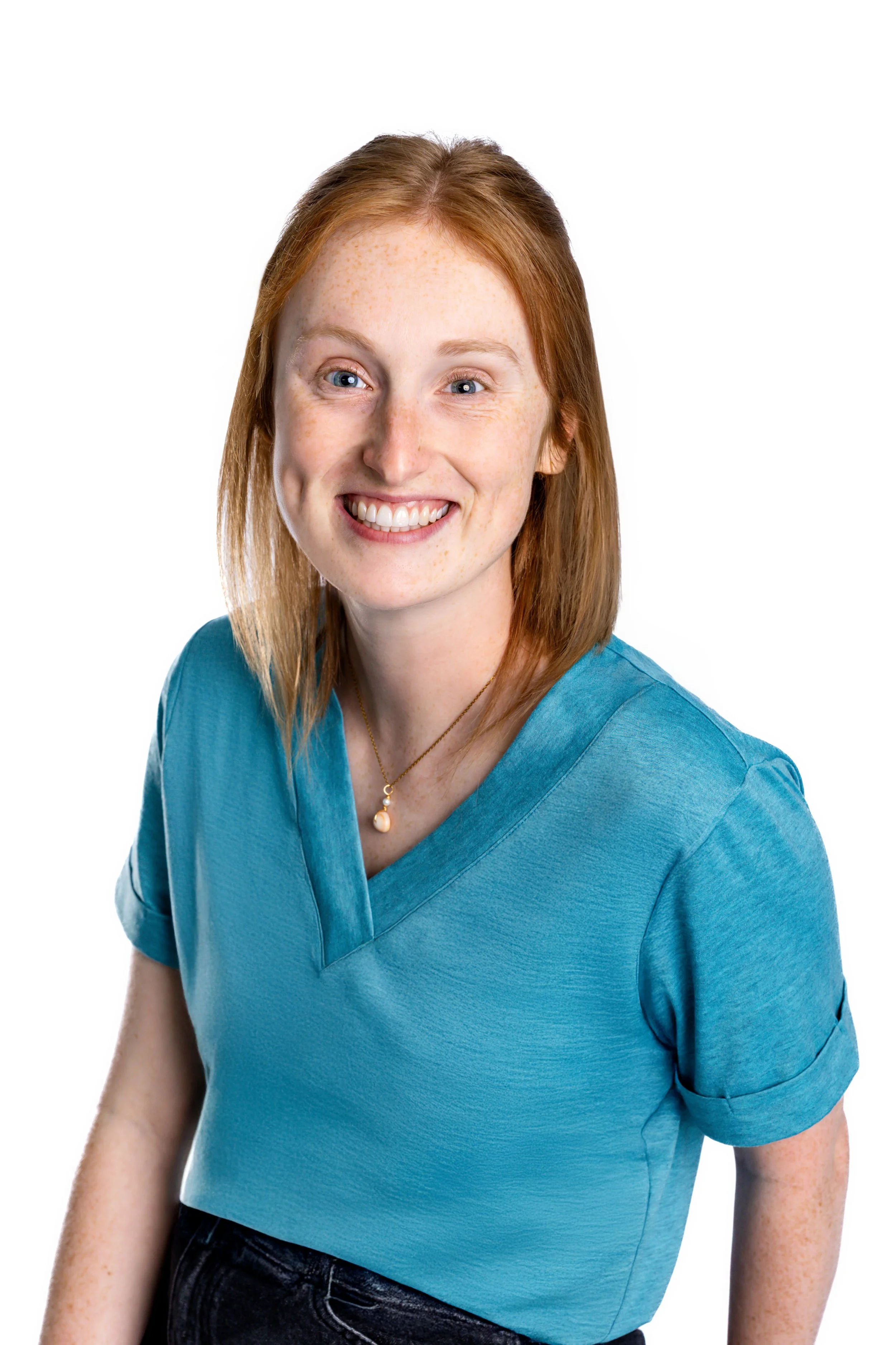 A woman with red hair and fair skin sitting on a cream-colored armchair in a room with blue walls and furniture. She is smiling, wearing a teal shirt, black jeans, and beige sandals. To her left, on a small pink and blue side table, there is a lamp with a woven base and a framed sign that reads "GO WITH THE WAVES." In the background, there is a blue cabinet with flowers on top and walls decorated with blue-framed windows.