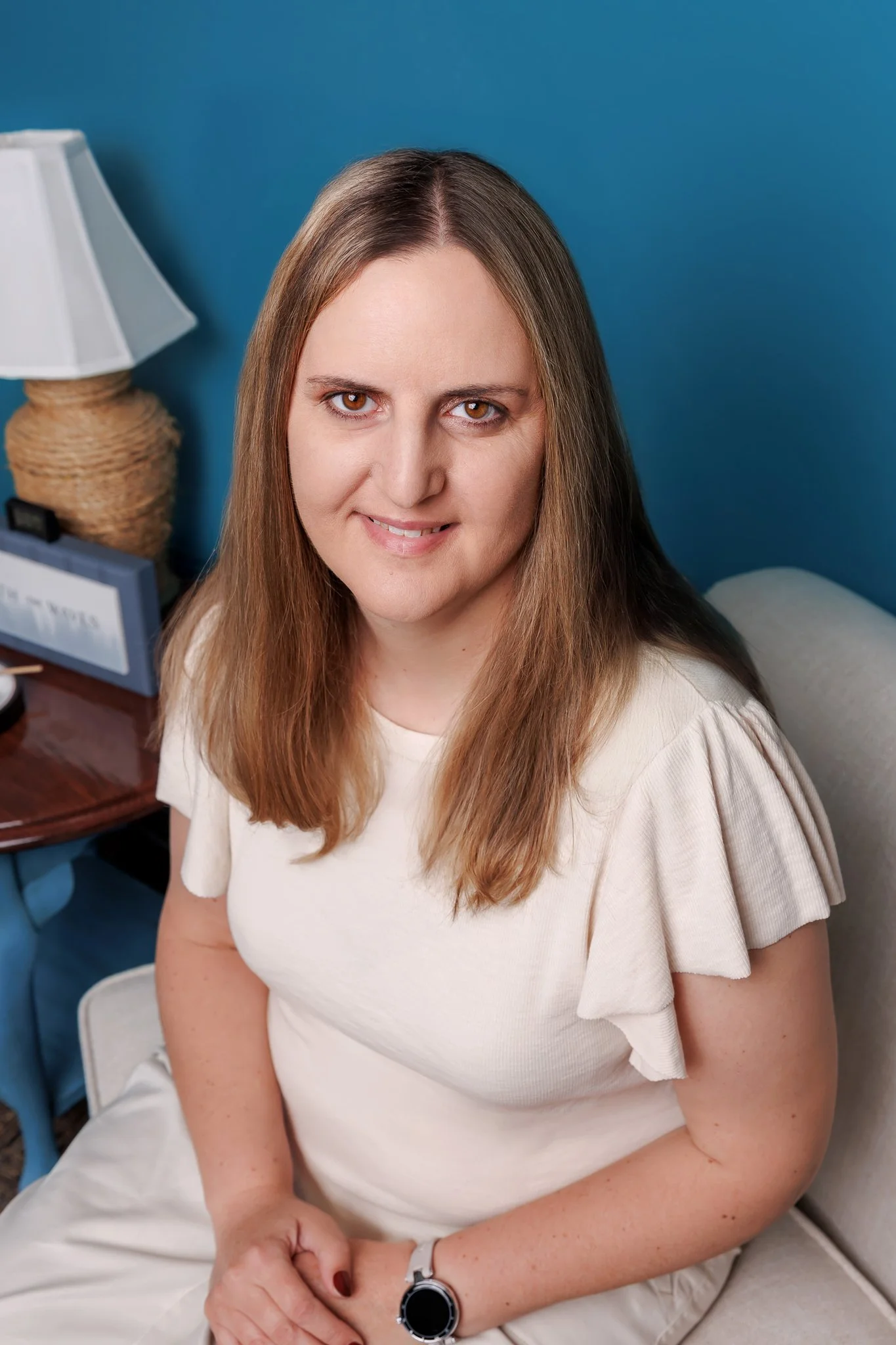 A woman with long brown hair and brown eyes sitting on a bed, wearing a white short-sleeved top, with a blue background, a white bedside lamp, and a nightstand with a sign and other items.