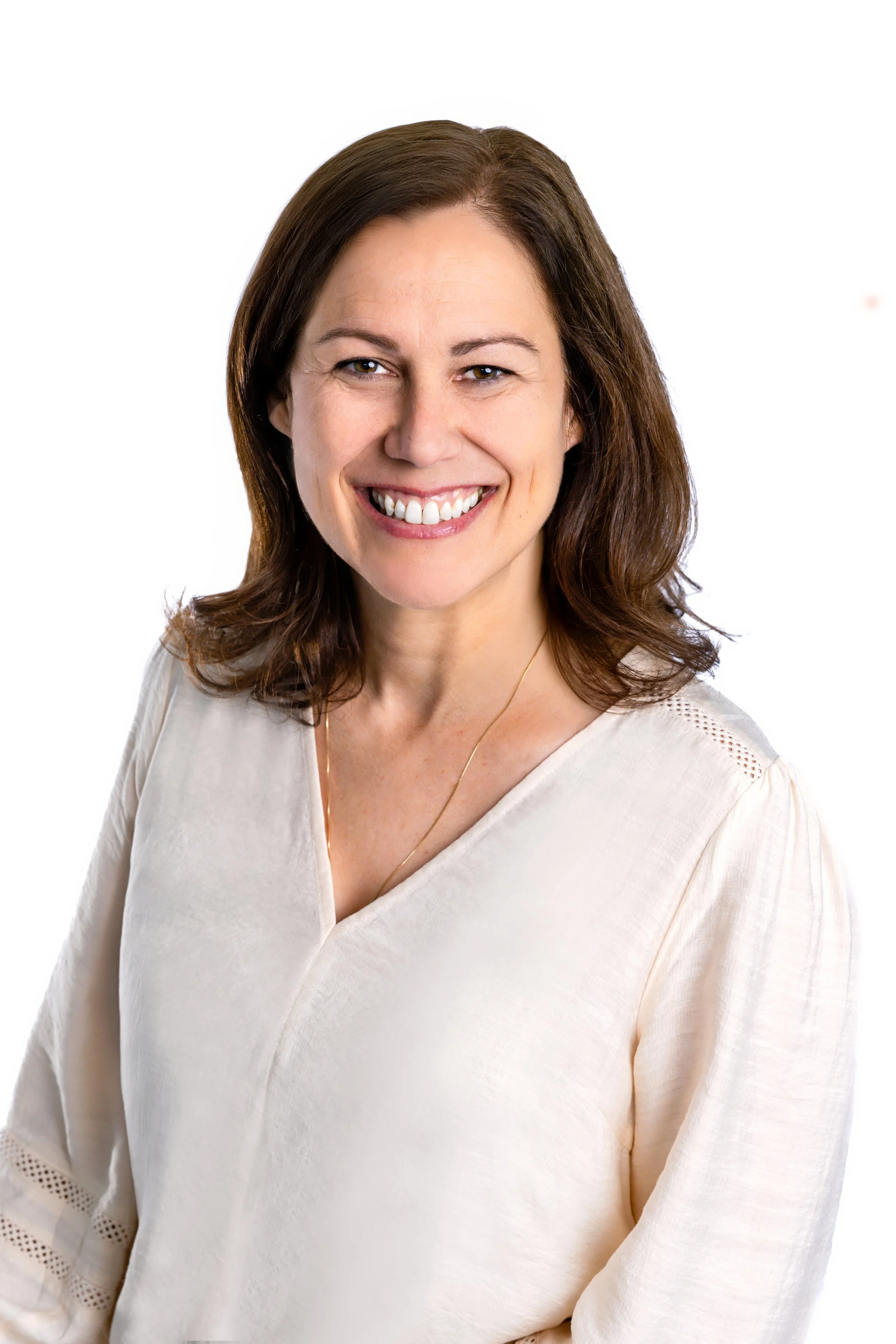 A woman with shoulder-length brown hair and a wide smile, wearing a cream-colored blouse and wide-legged pants, sitting on a beige armchair in a room with light blue walls. To her left is a small round wooden side table with a lamp and a sign that says "Go with the Waves." Behind her, there is a tall floor lamp with a white shade, and a blue dresser with some flowers on top. There are two wall-mounted blue framed mirrors or panels on the wall.