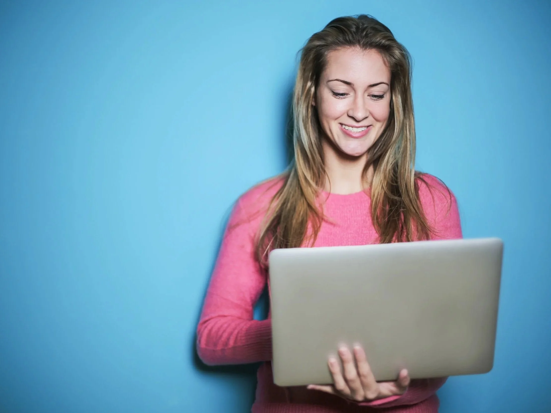 A woman holding and looking at a laptop, smiling, against a blue background.