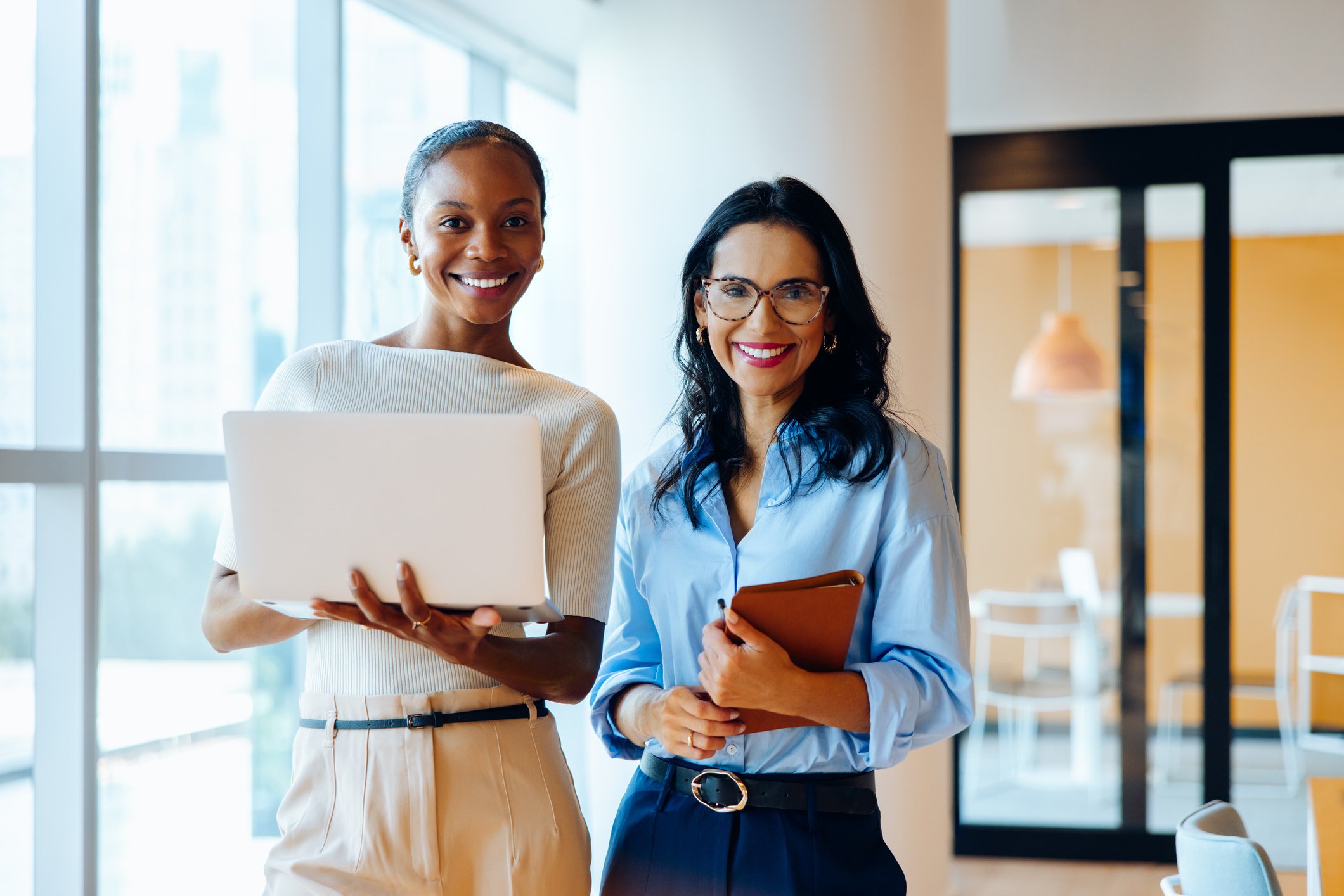Two women standing against a plain wall, one holding a pointer and the other holding a folder.