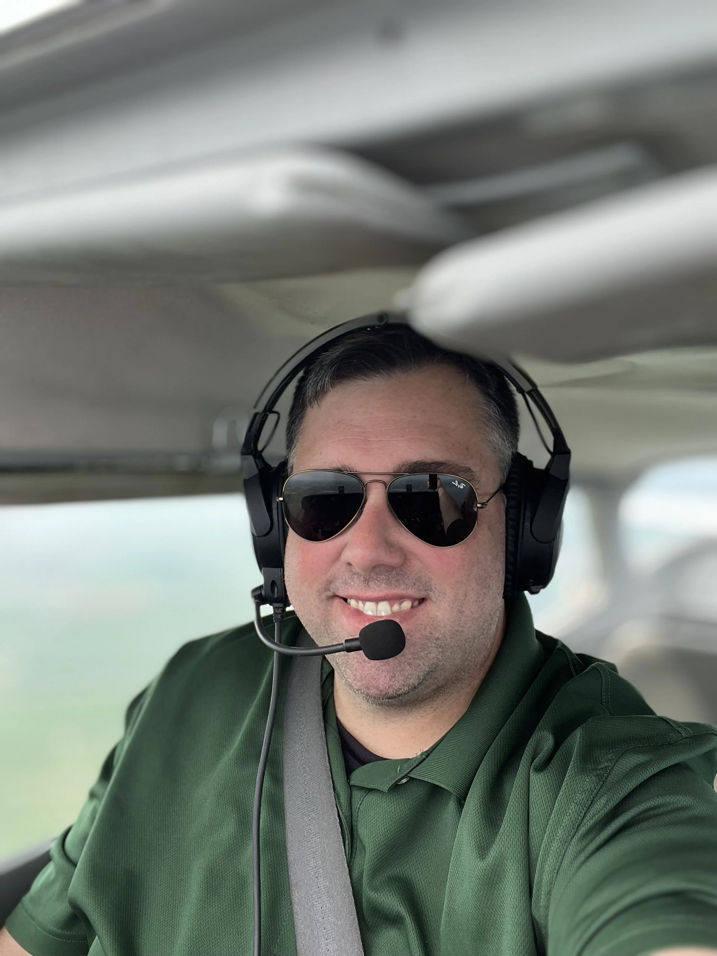 Man wearing sunglasses, headset, and green jacket smiling inside a small aircraft or cockpit.
