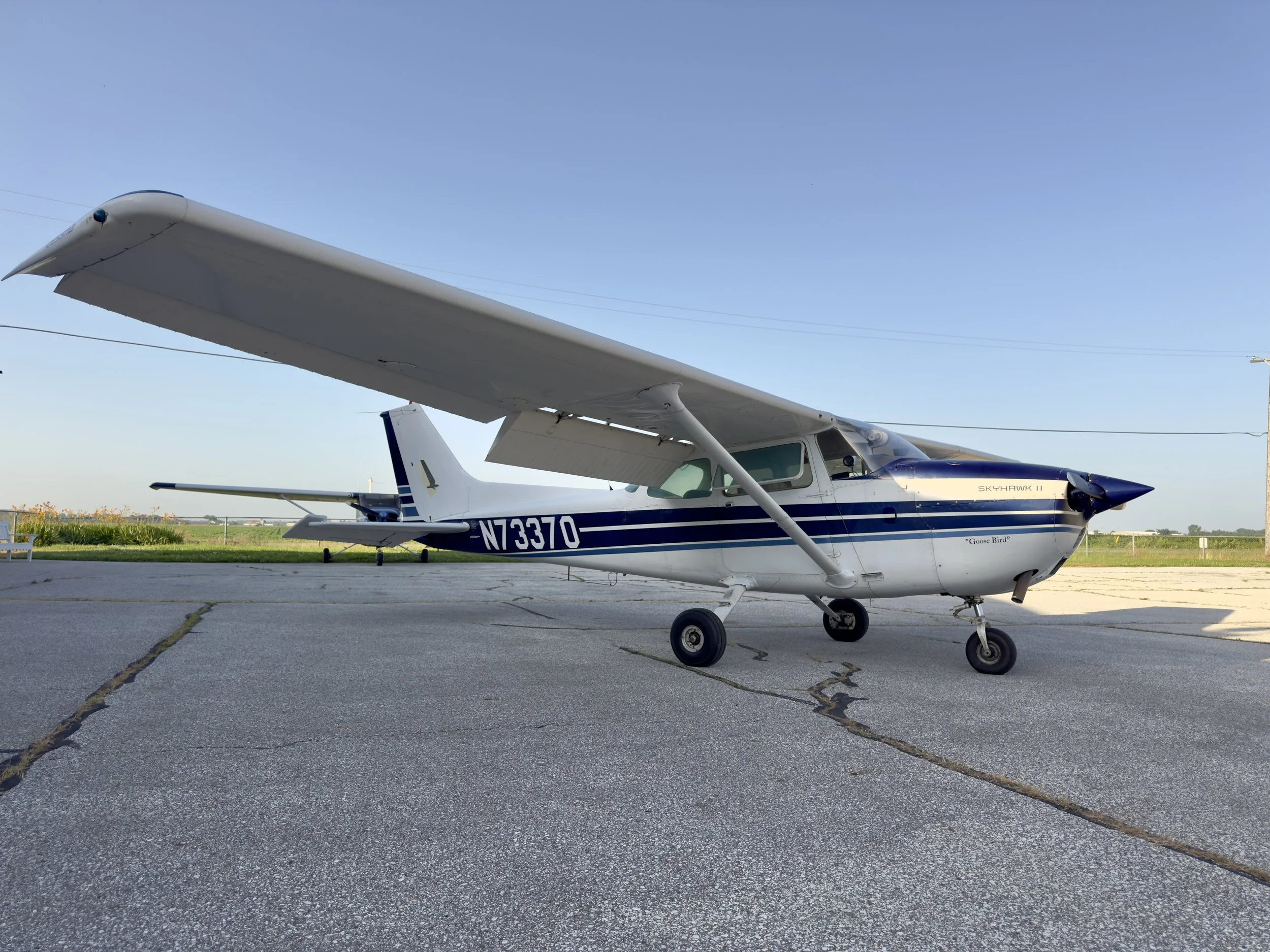 Small white and blue propeller airplane on a concrete tarmac with grass and some buildings in the distance, under a clear blue sky.
