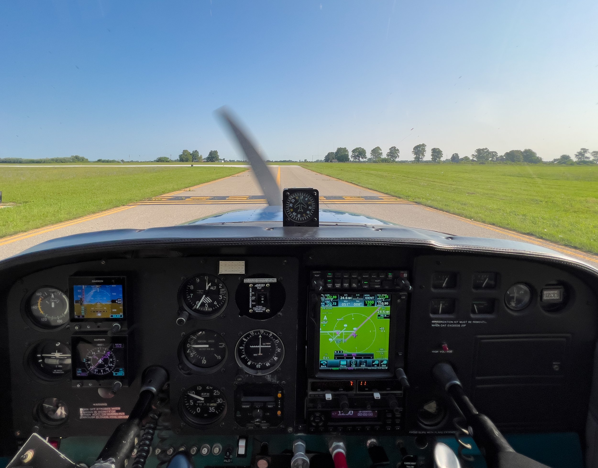 Pilot cockpit view showing various gauges, screens, and controls, with a view of a flat landscape with green fields and trees through the windshield.