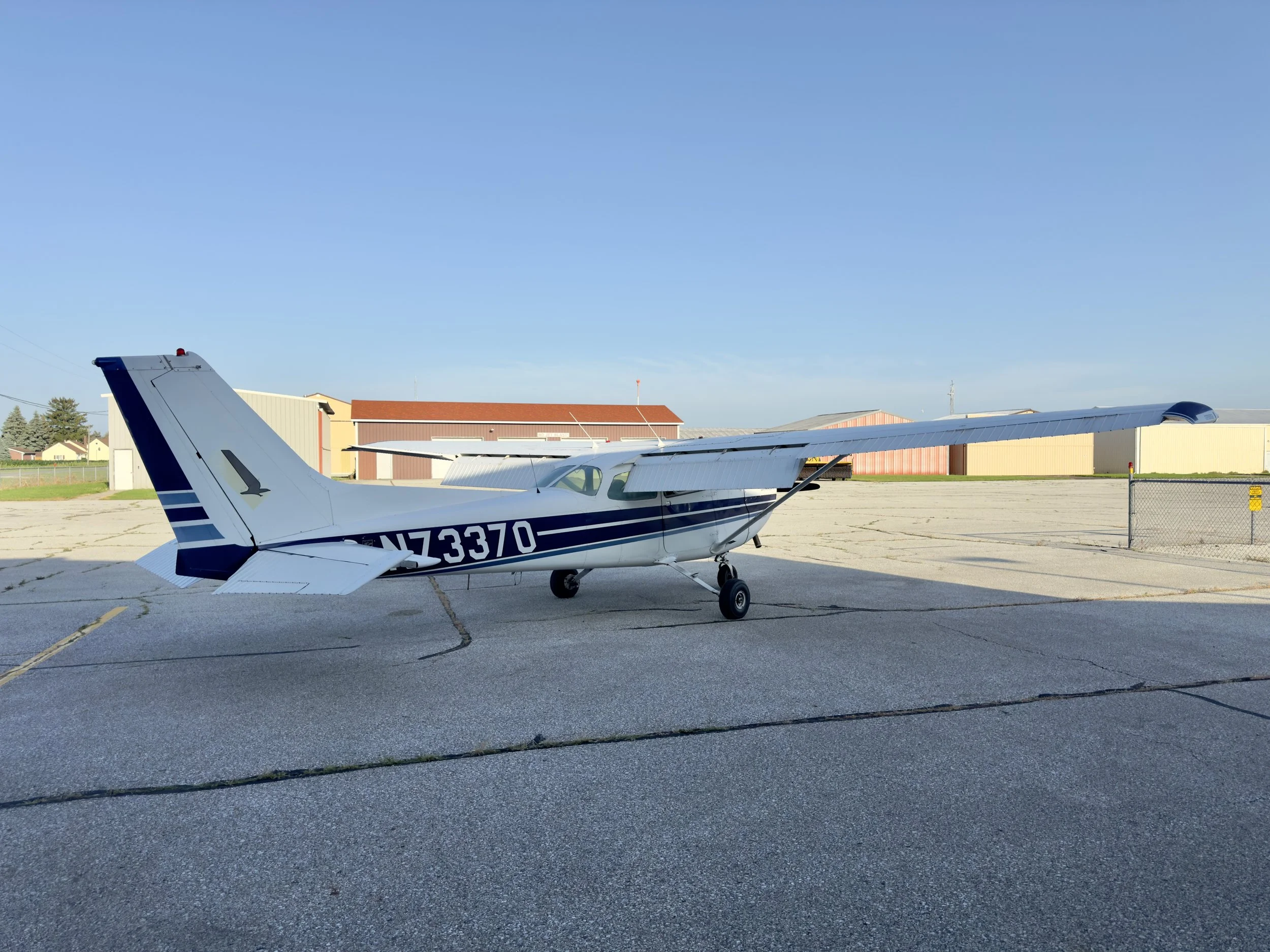 Small white and blue Cessna airplane parked on a runway with some hangars and buildings in the background under a clear blue sky.