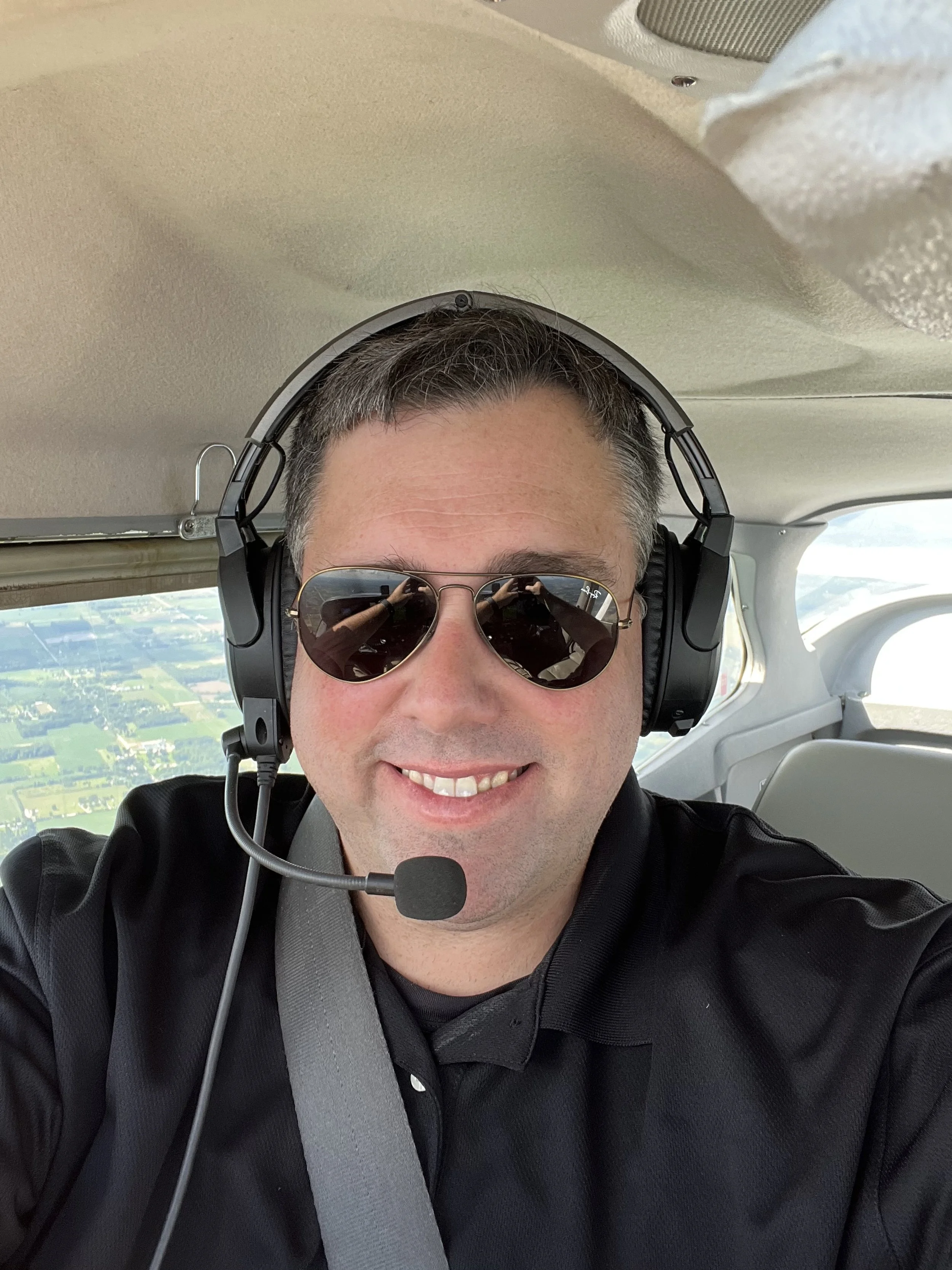 A man wearing sunglasses, a headset with microphone, and a black shirt sitting in the cockpit of an airplane with a view of the sky, clouds, and landscape below through the windows.