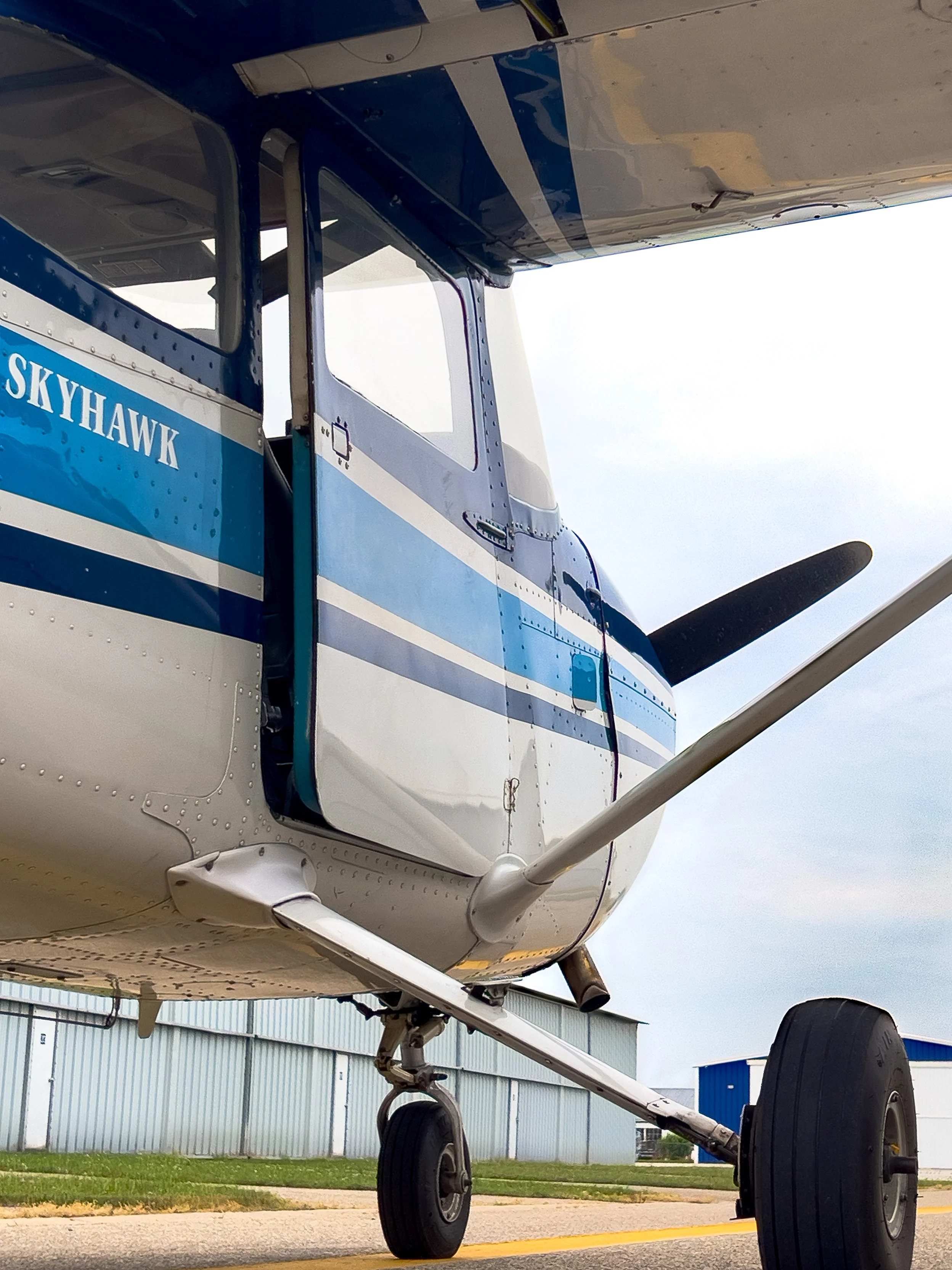 Close-up of the front part of a white and blue SkyHawk aircraft, focusing on the wheel and landing gear, with a hangar and blue sky in the background.