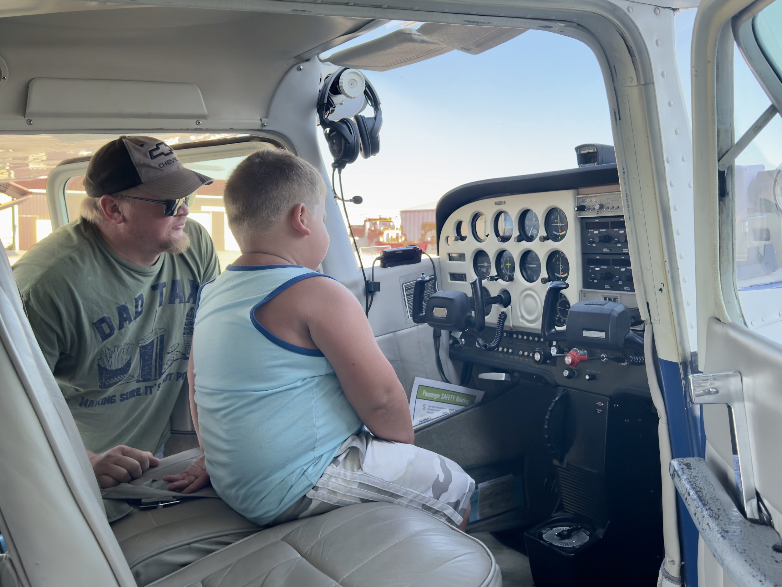 A man and a young boy sitting inside a small aircraft cockpit, with the man leaning towards the boy, possibly explaining flight instruments, on a sunny day.