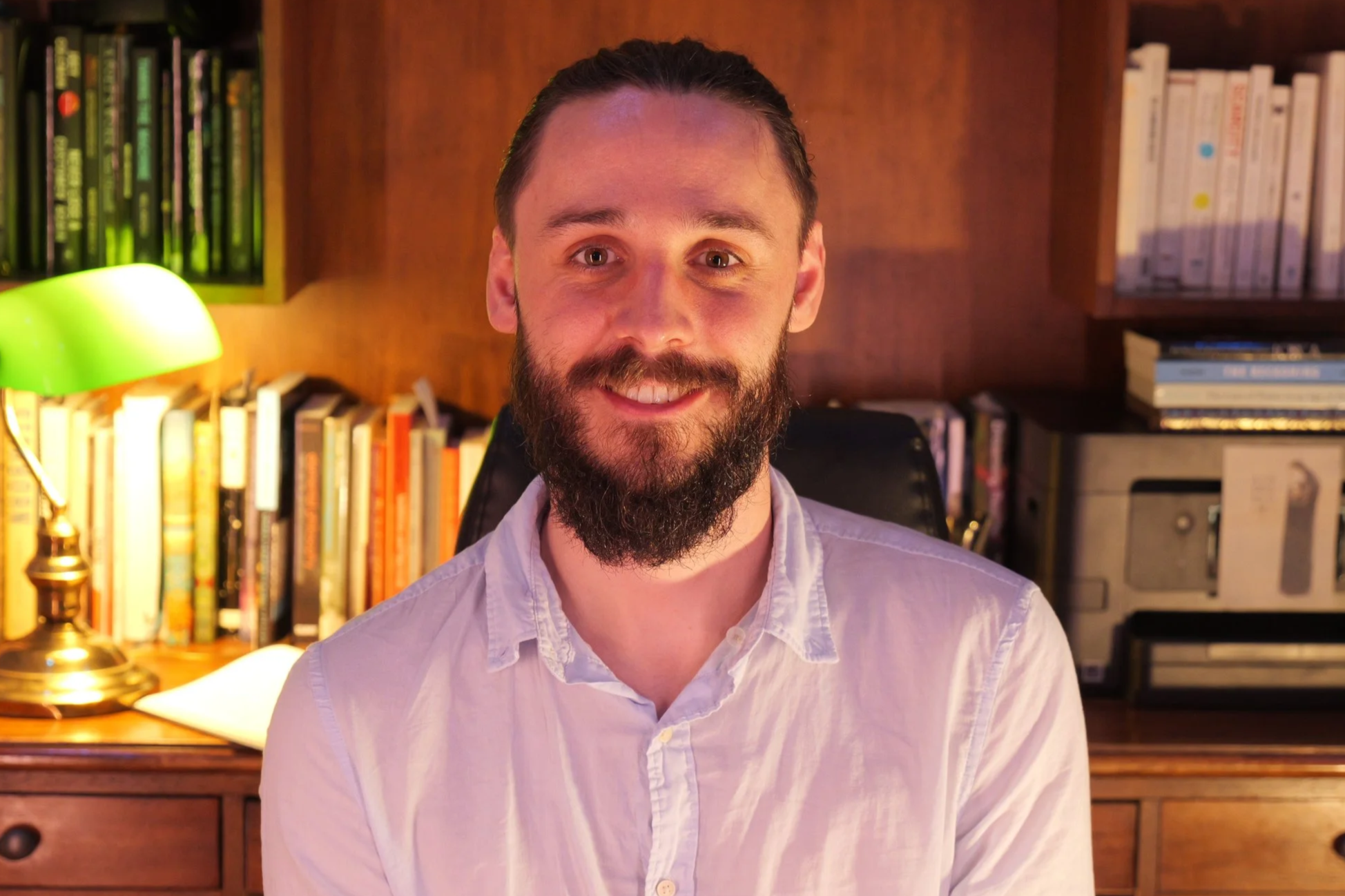 Photo of Malibu Poet Laureate and Founder of The Poetry Vessel, Nathan Hassall. Shown sitting in his home office with wooden furniture and bookshelves, illuminated by a green desk lamp.