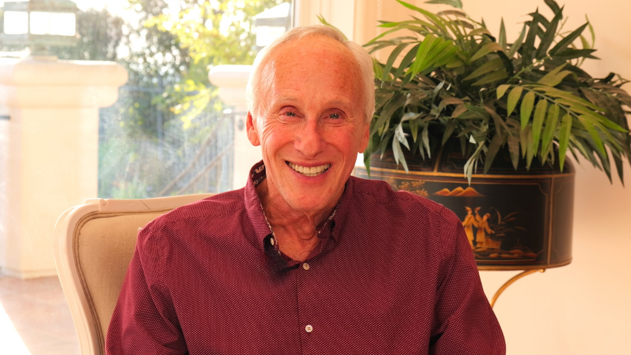 Photo of Neuroscience Researcher and Best-selling Author of "How Words Change Your Brain" Mark Waldman. Sitting in a beige upholstered armchair in a sunlit room with large window and a decorative plant in a black vase on a stand to his right.
