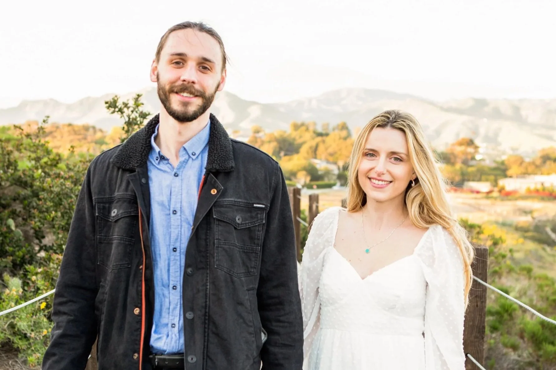 A man and woman stand outdoors in a scenic landscape with hills and trees in the background during daylight. The man has a beard and is wearing a black jacket over a blue shirt. The woman has long blonde hair and is wearing a white dress.