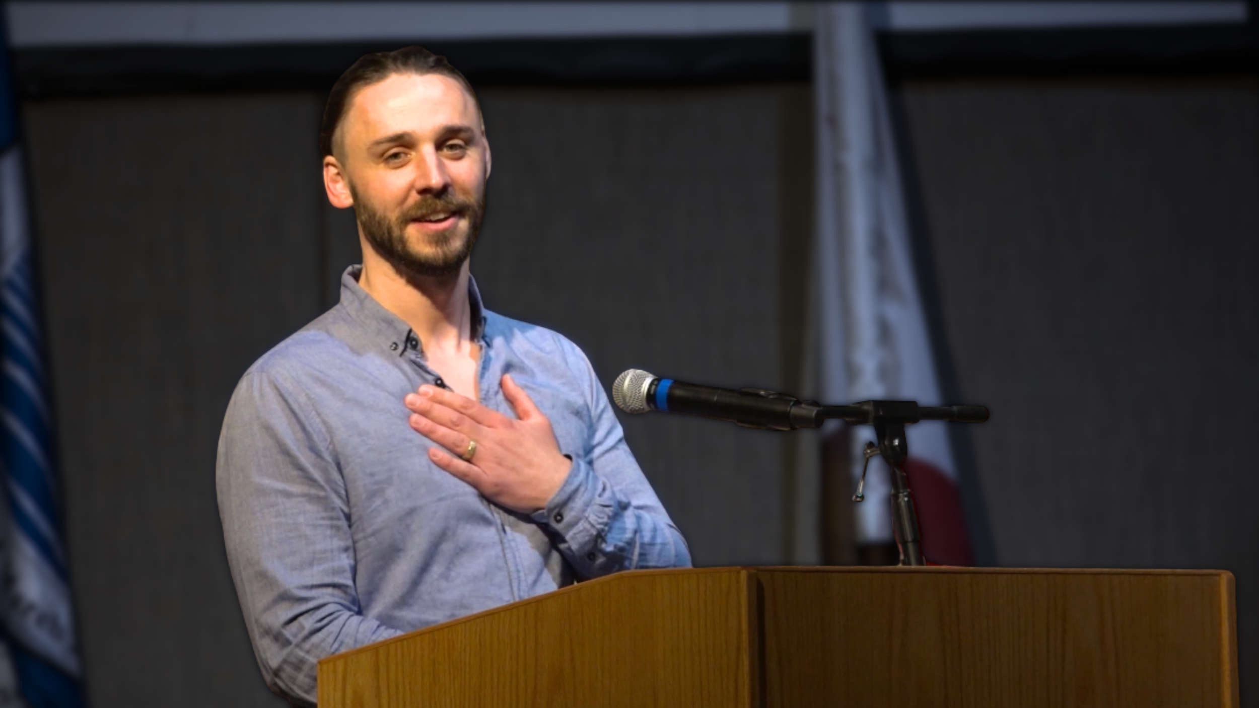 A man with a beard and short hair, wearing a gray shirt, standing at a podium with his right hand over his chest, speaking into a microphone.
