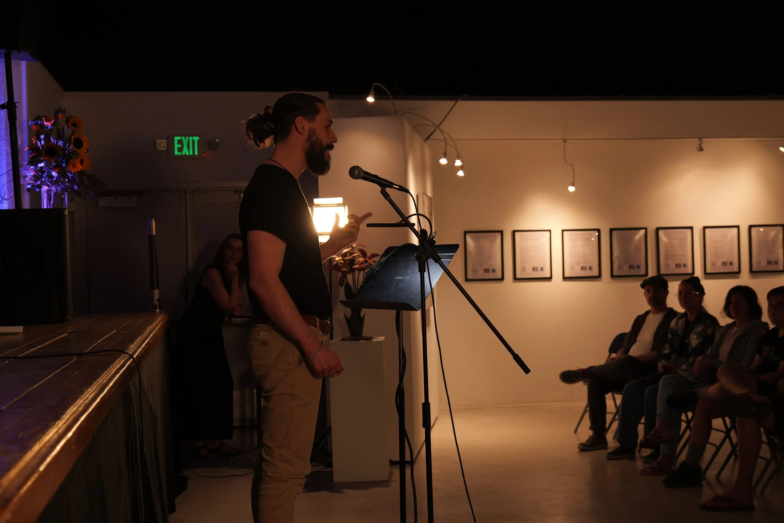 A man with long hair and a beard speaking into a microphone at an indoor event, with an audience seated and a woman smiling in the background.