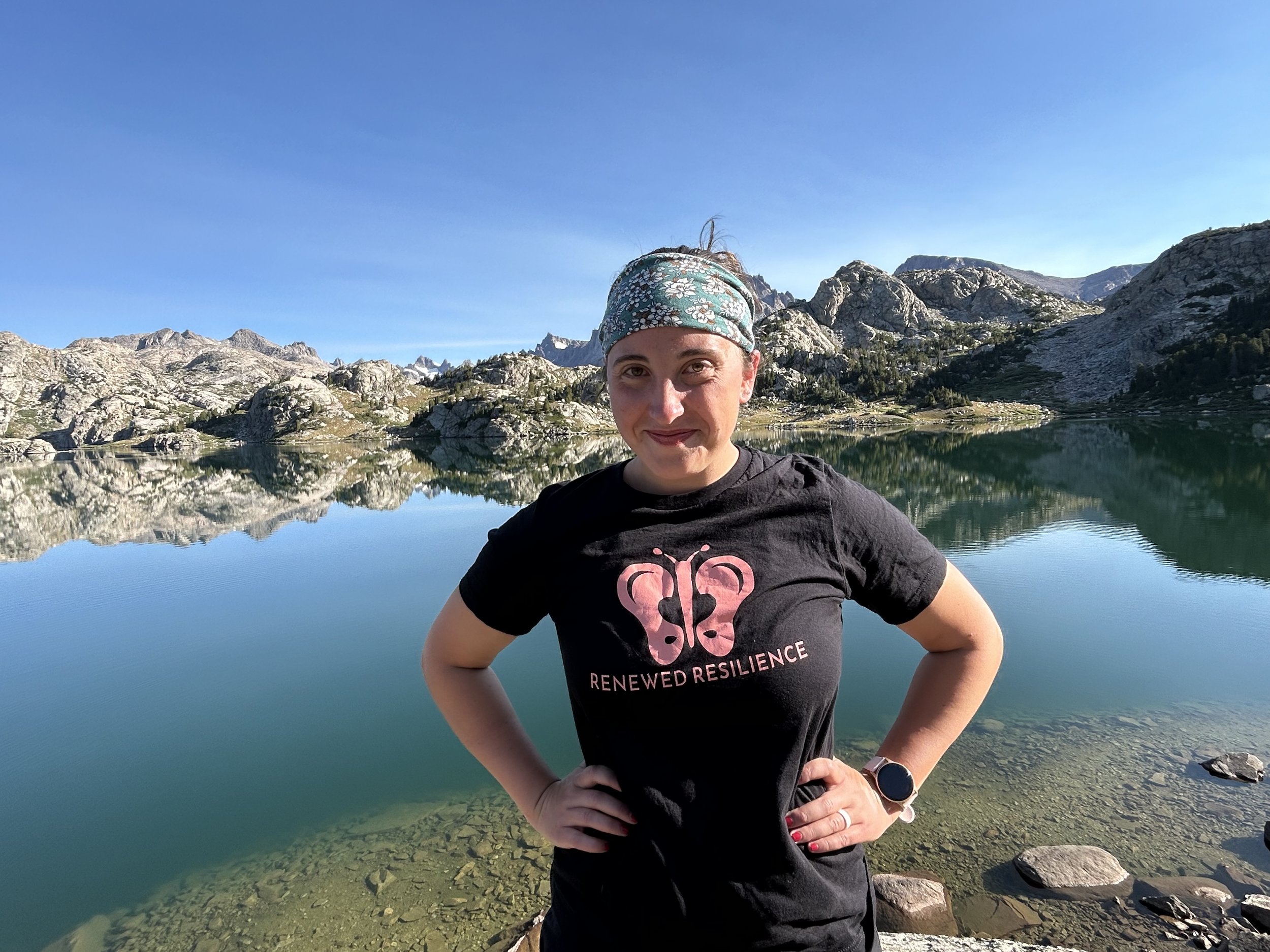 Person posing near a mountain lake with rocky hills and clear blue sky in the background.