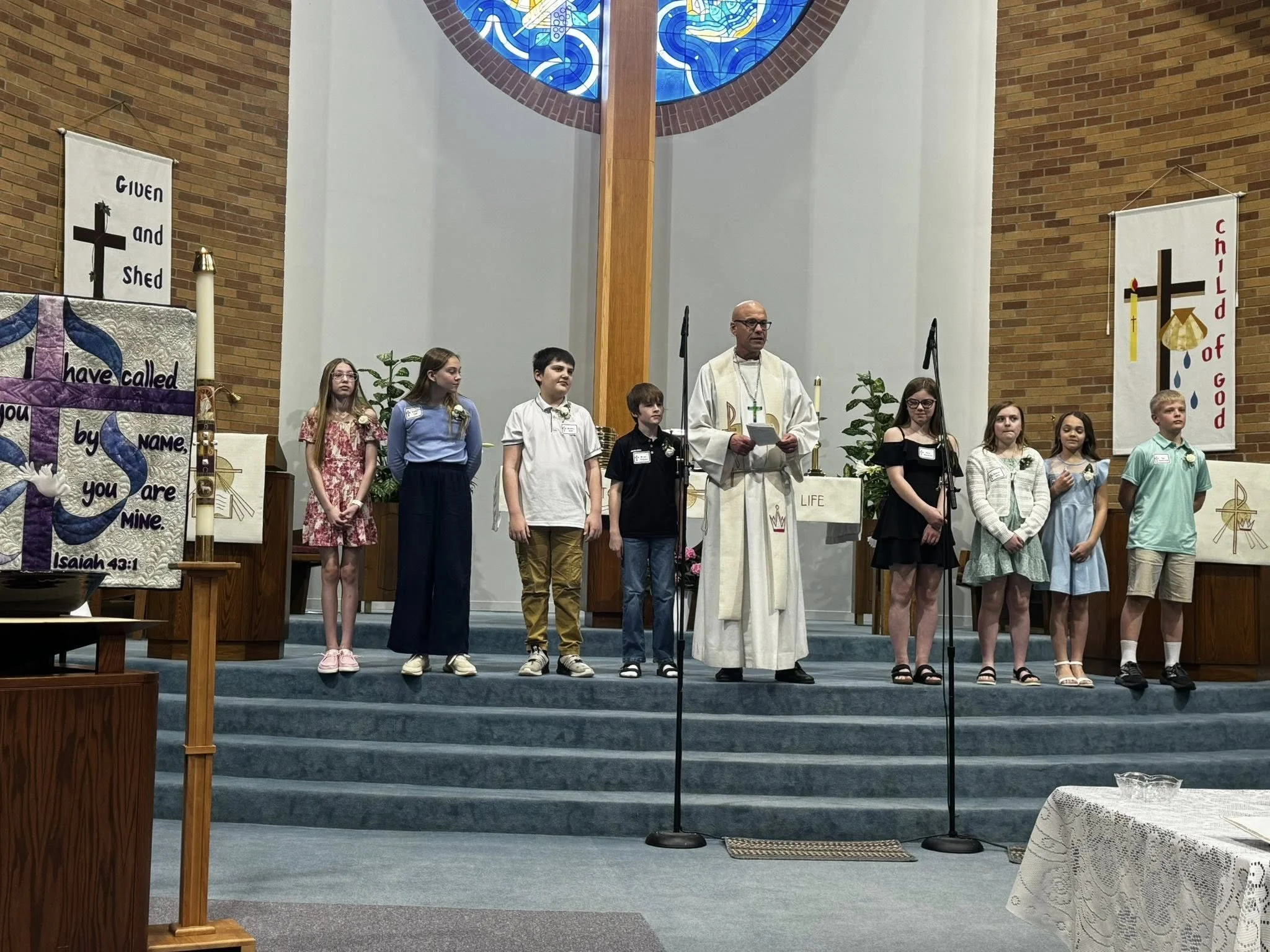 Children standing on stage in church, with a minister or priest, during a religious service or ceremony, with banners and decorations in the background.