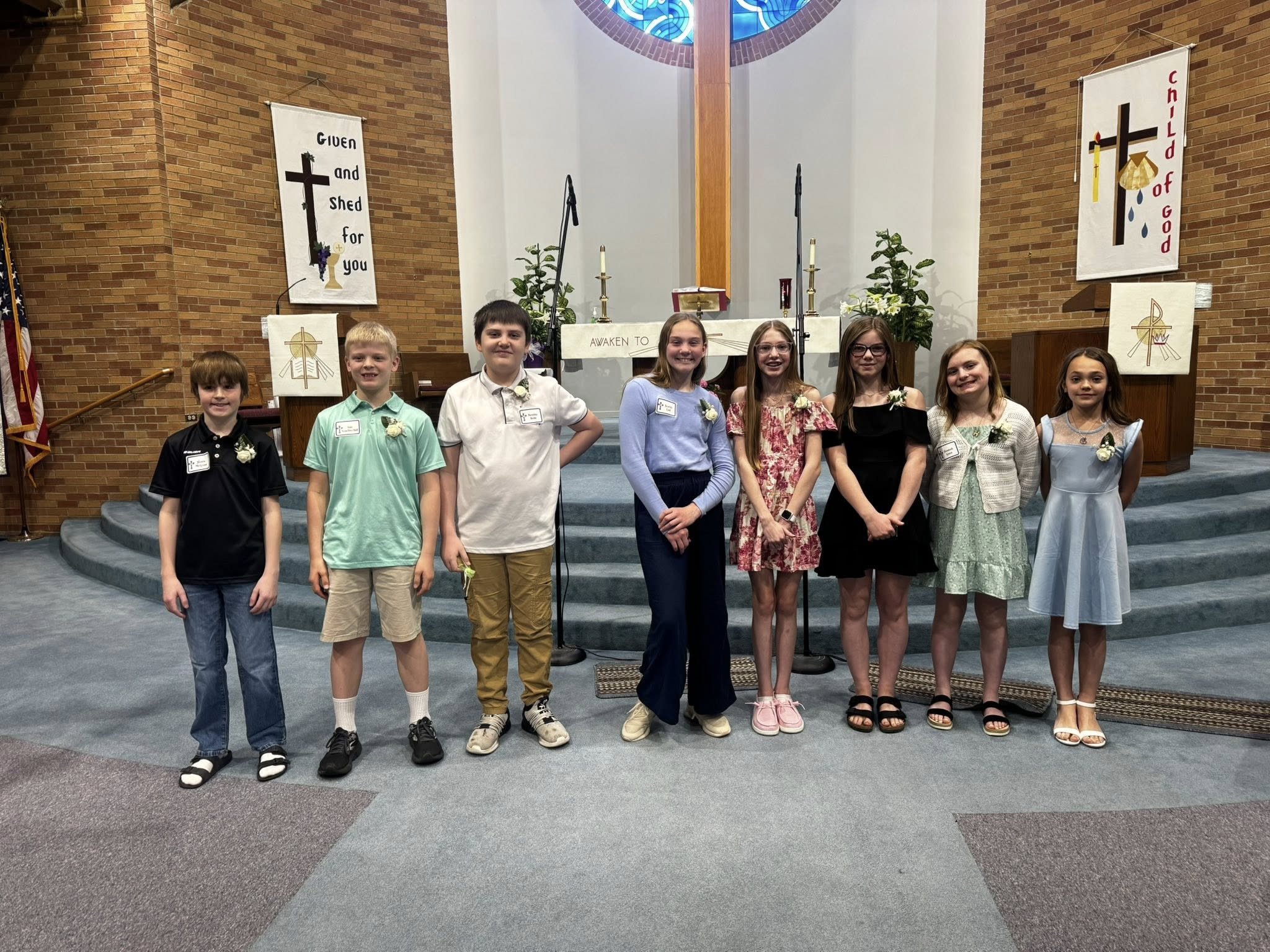 A group of nine children standing on the steps inside a church, dressed in semi-formal attire, with a brick wall, banners, and religious symbols in the background.