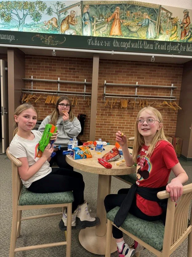 Three young girls sitting around a circular table in a room with brick wall and coat hangers. They are eating snacks and playing with toy water guns. Two of the girls have glasses, and one has a red T-shirt. Snacks and water bottles are on the table, and a mural or painted mural is above the brick wall.
