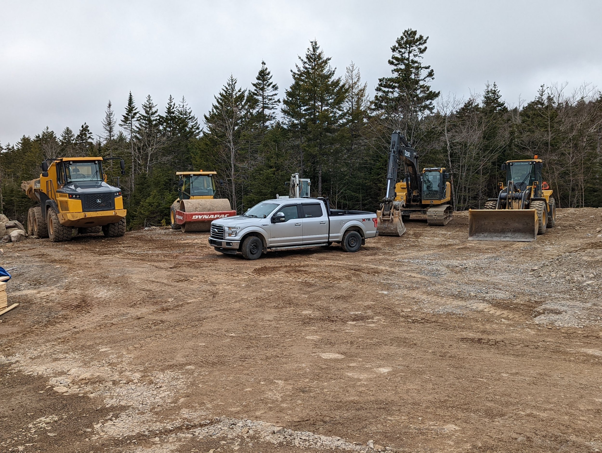 Construction site with heavy machinery and a pickup truck, surrounded by trees.