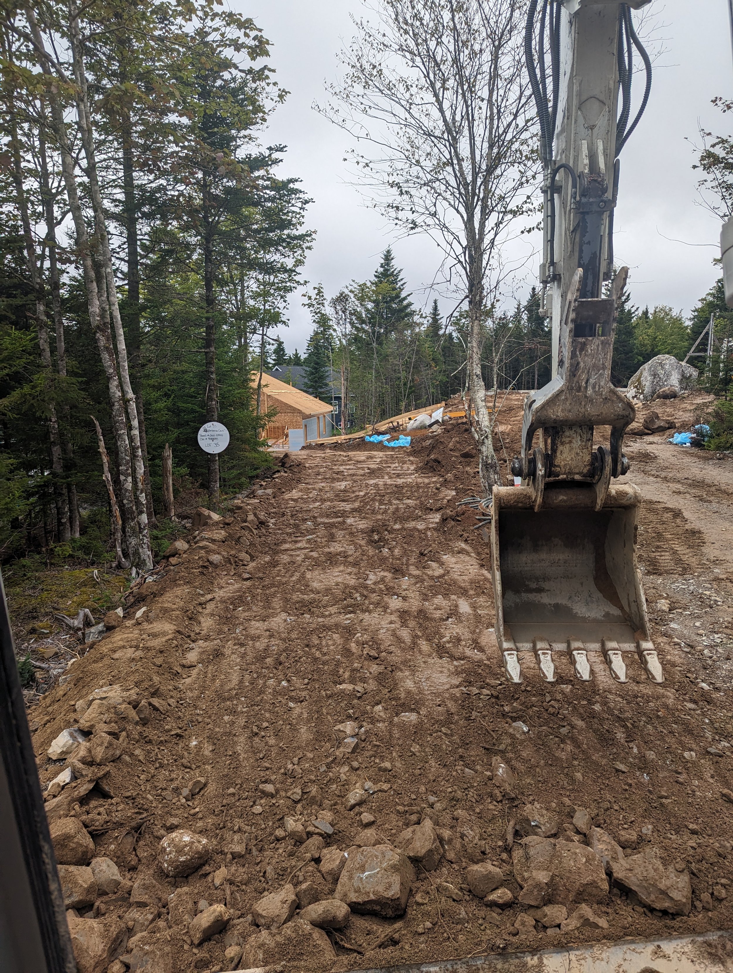 Construction site with dirt path and excavator bucket, surrounded by trees, leading to a partially built wooden structure.