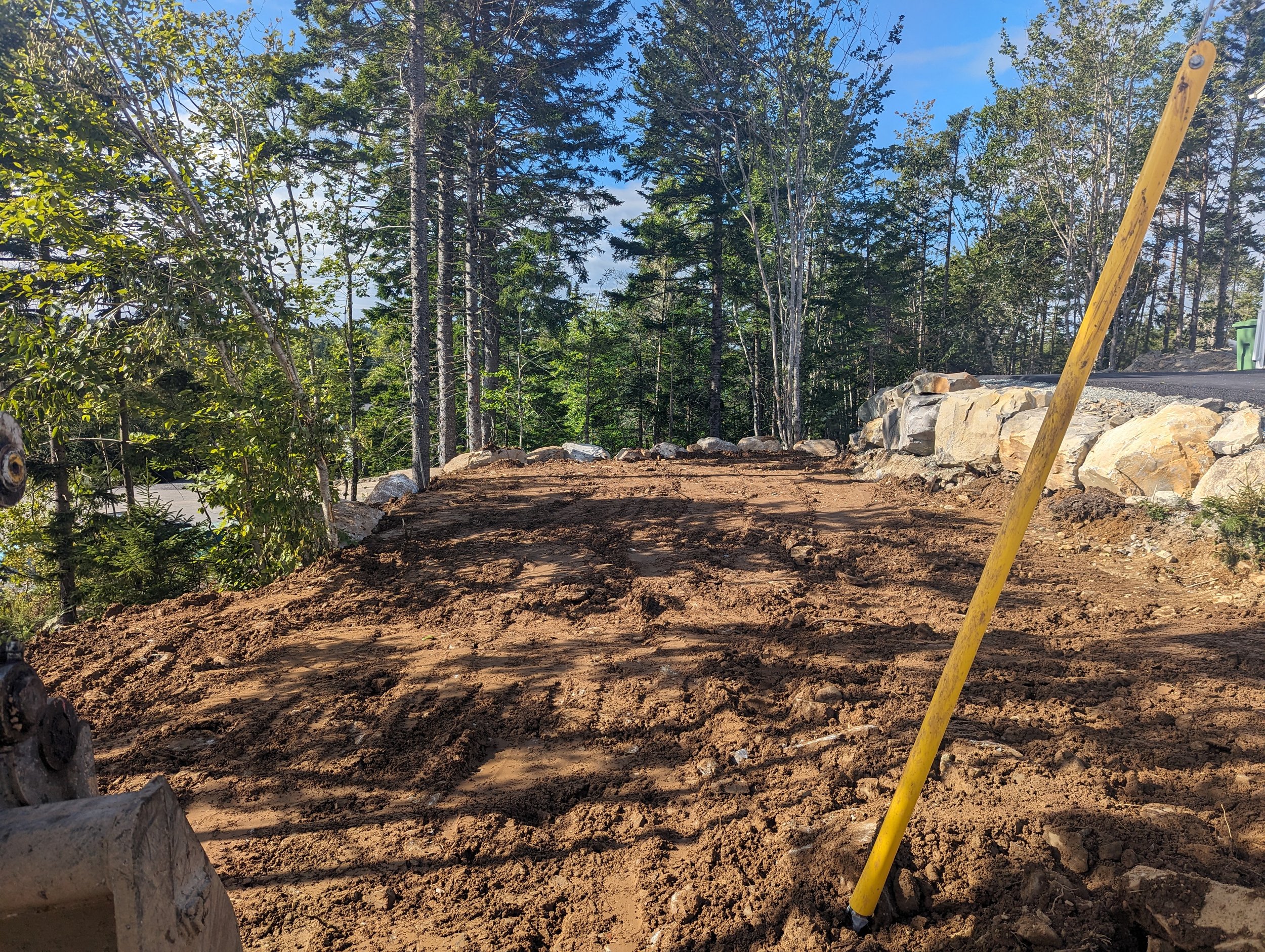 Cleared dirt area surrounded by trees, with a yellow pole in the foreground, and a rocky edge separating the dirt from a paved road.