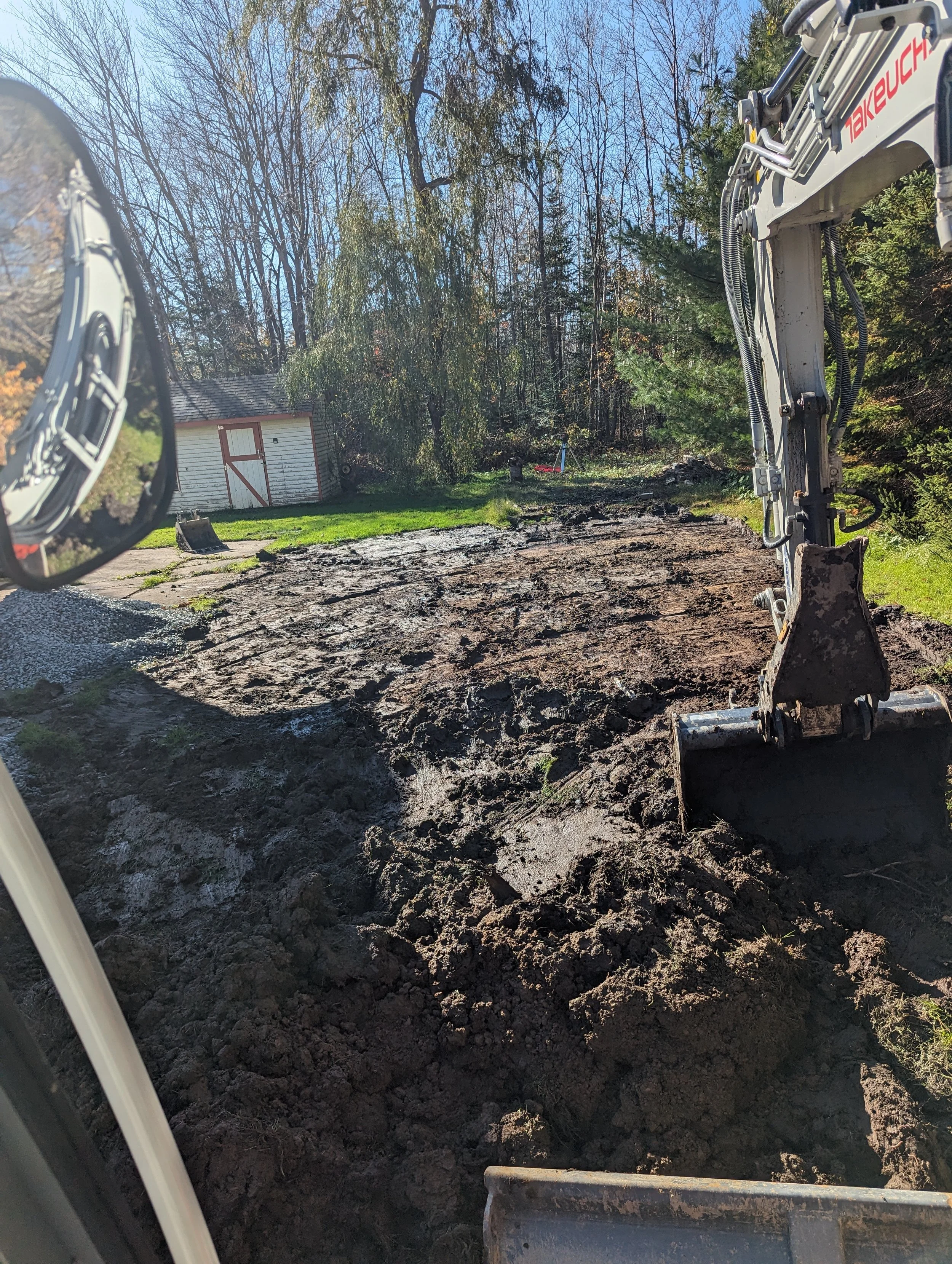Excavator digging in a muddy yard with trees and a small white shed in the background on a sunny day.