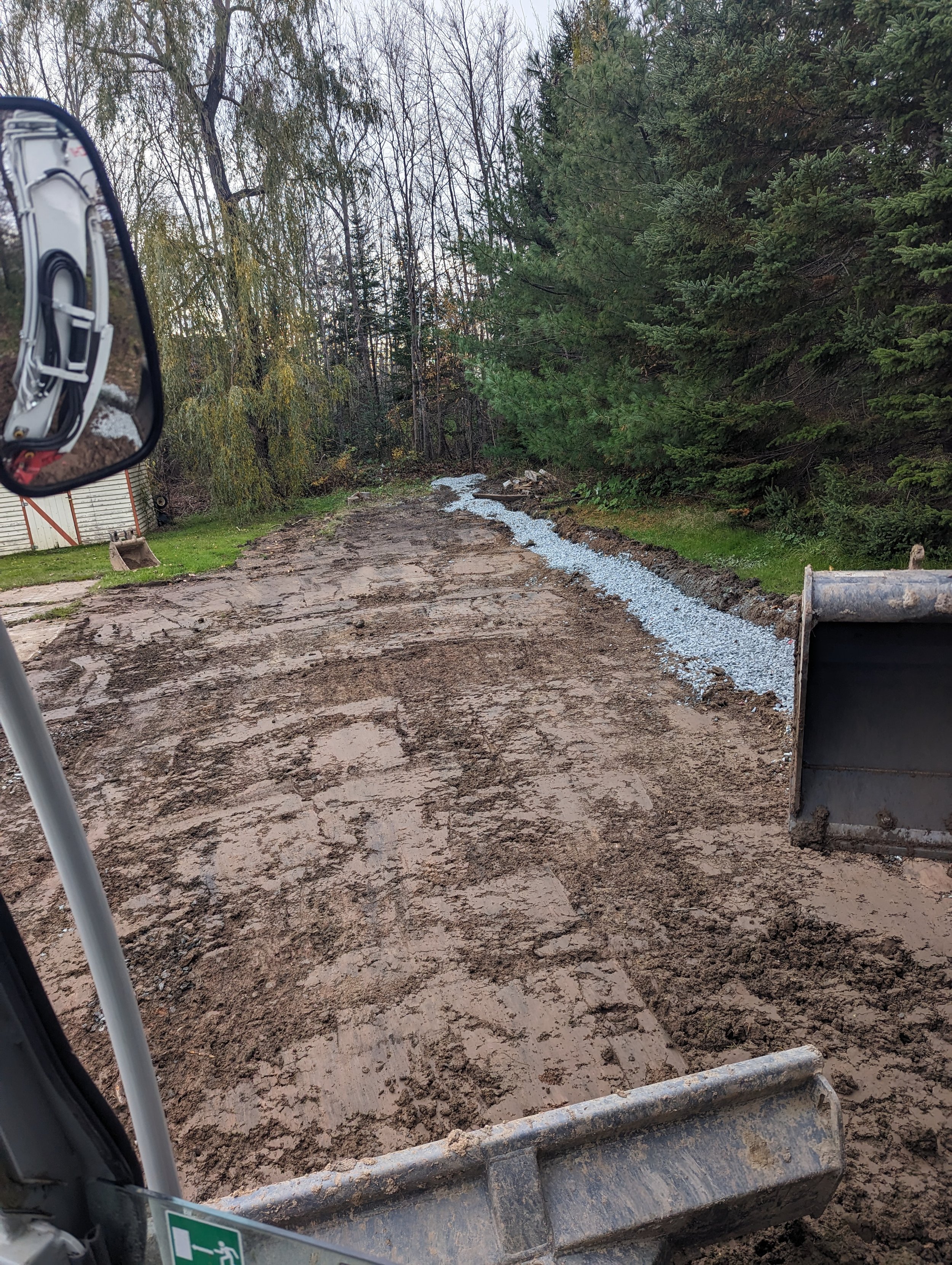 Construction site with muddy ground and a bulldozer blade; gravel spread along the side, surrounded by trees.