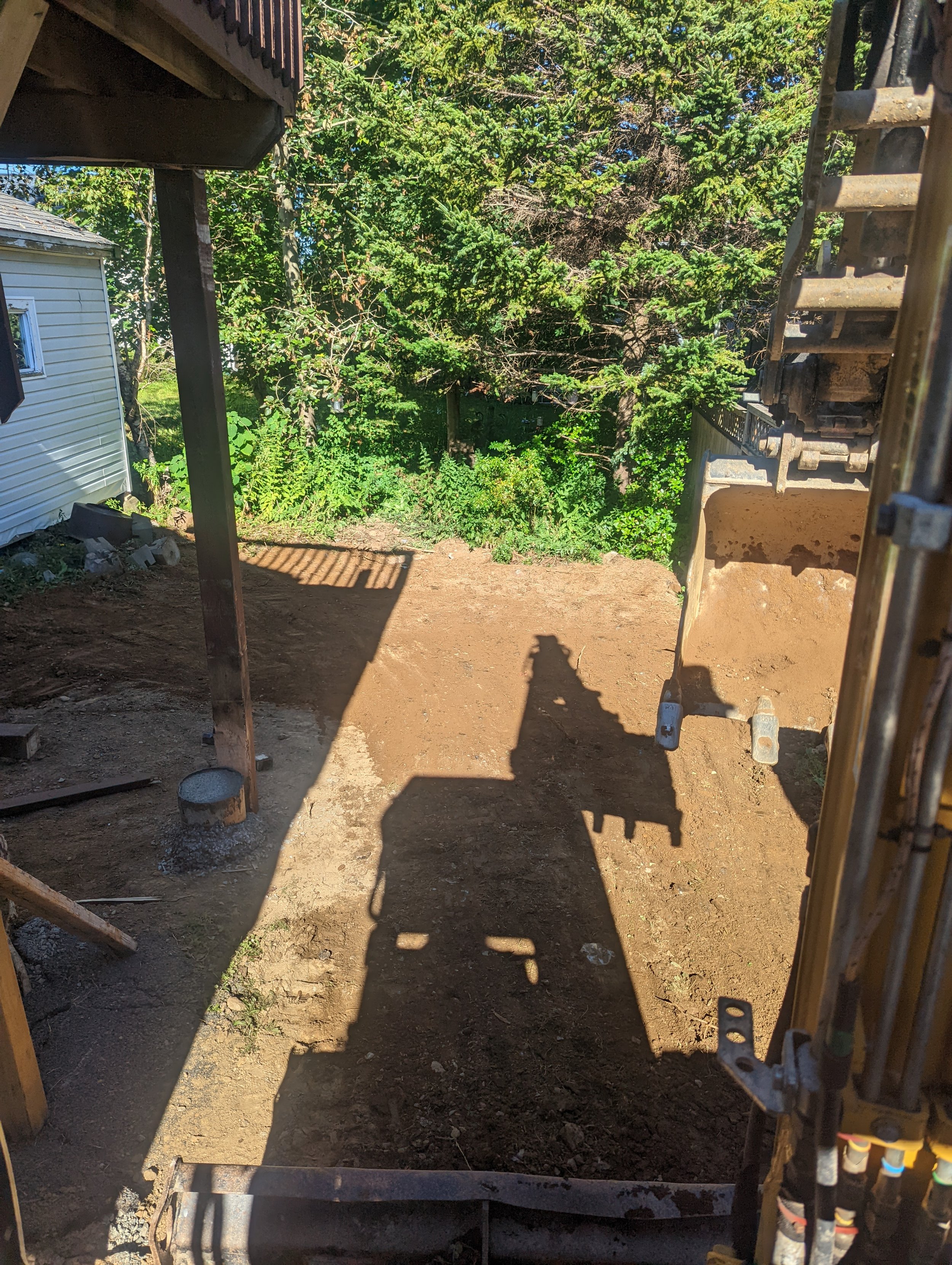 Excavation site with dirt clearing, trees in the background, a building on the left, and shadows from a construction vehicle's arm.