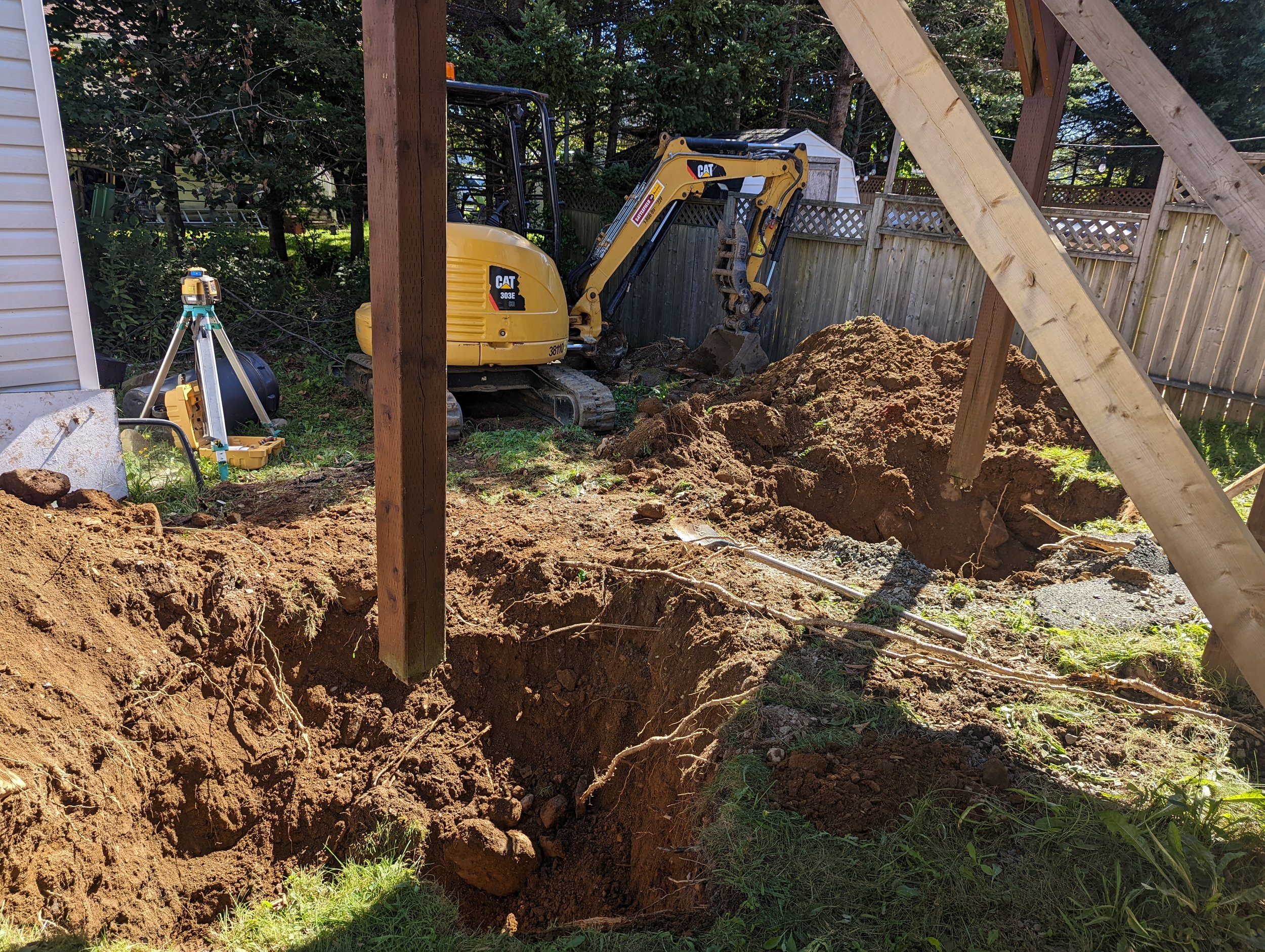 Excavation site with yellow mini excavator, wooden beams, and scattered dirt in a residential backyard.